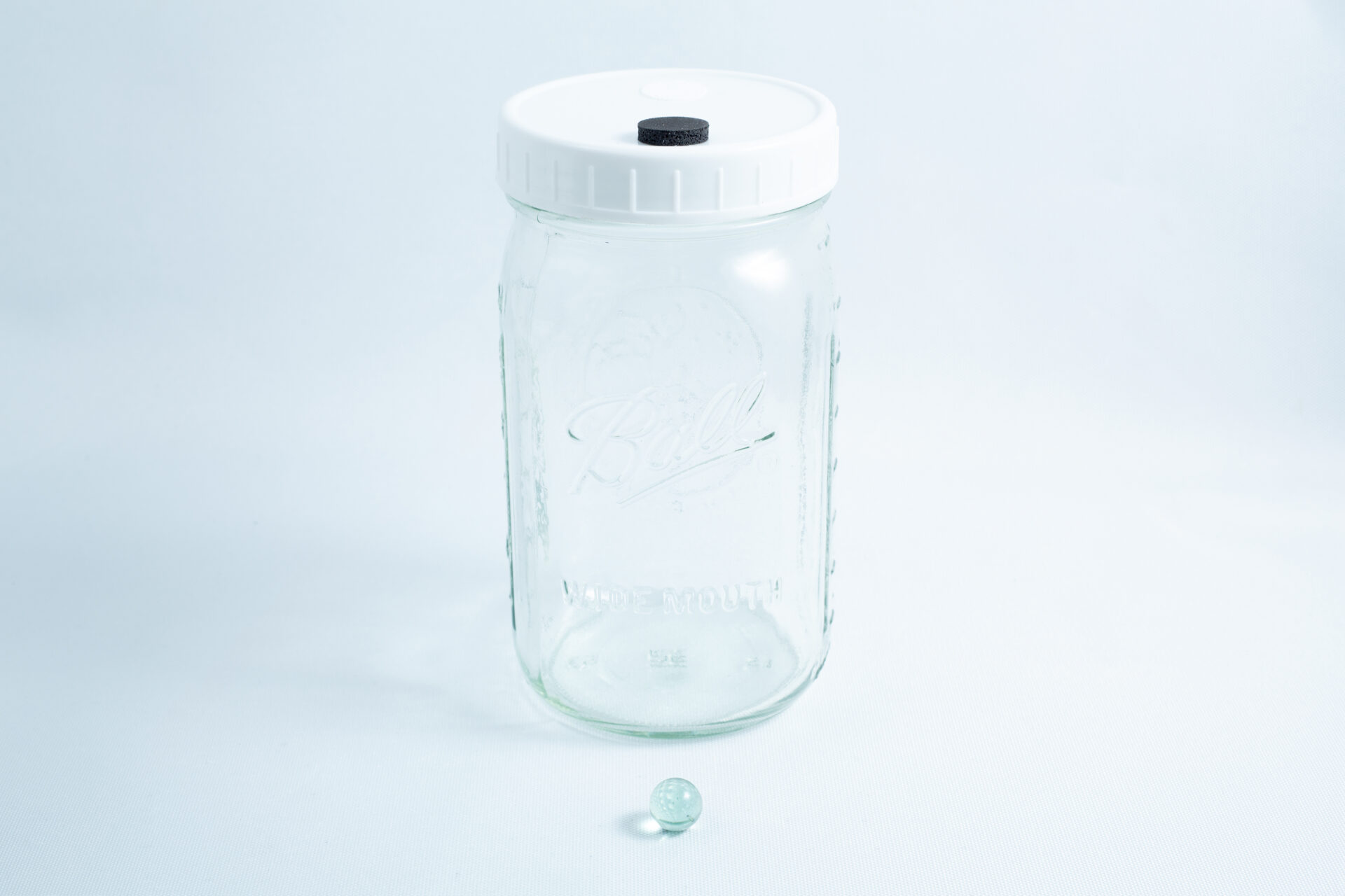 A Mycology Ball Kilner jar with injection port and air vent, 1L genuine and autoclavable, sits next to a small glass marble on a white background.