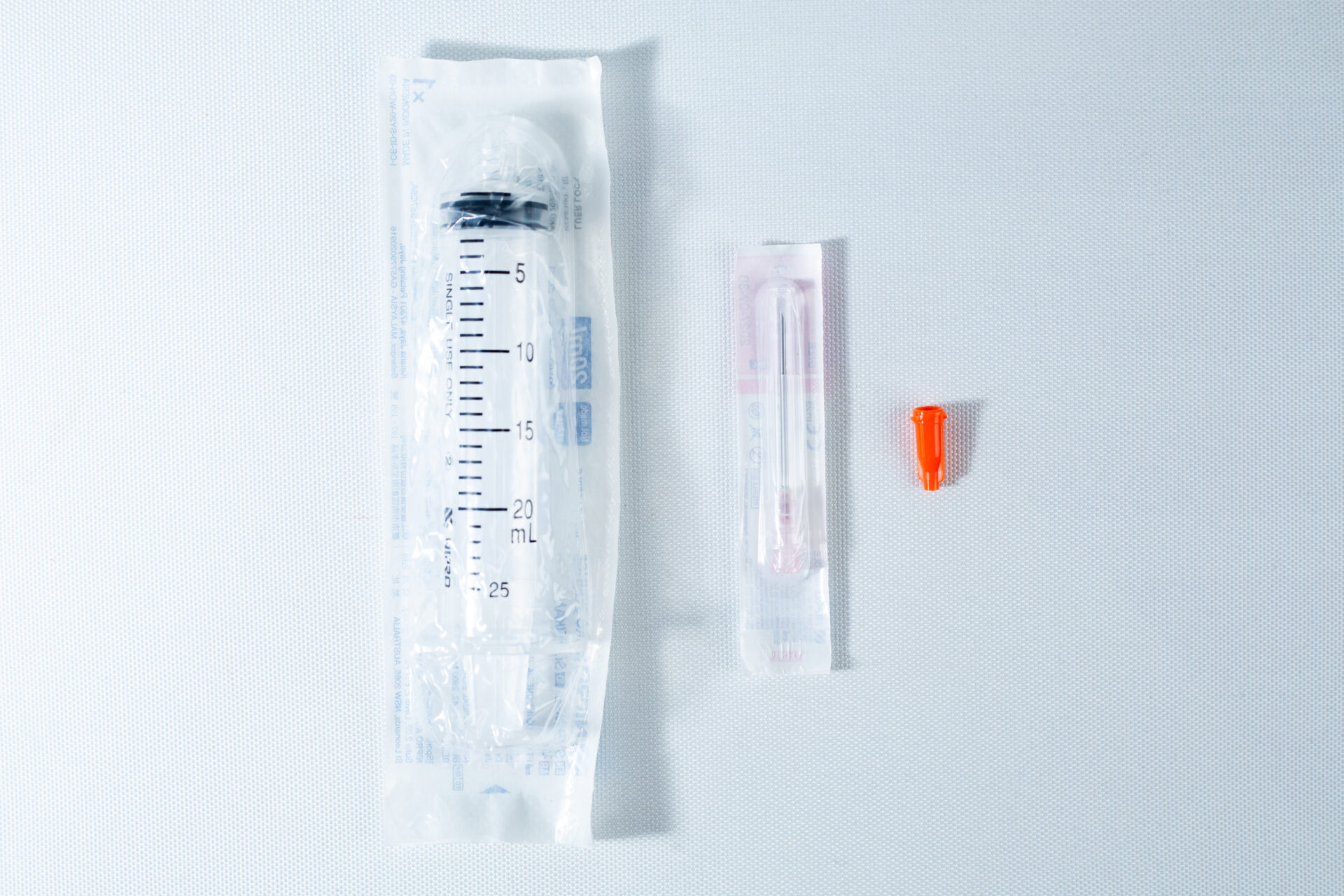 A packaged syringe, a packaged needle, and an orange needle cap on a white surface.