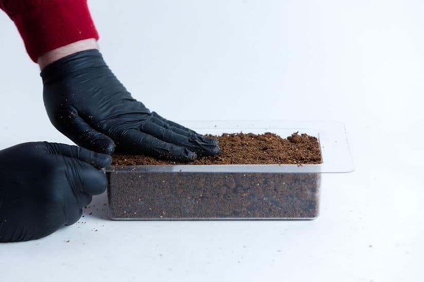 Hands in black gloves press soil into the Mycelium Mould Growth form - Brick (Reusable), shown in a clear plastic container against a white background.
