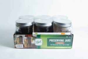 A set of six Ball Mason pint jars with wide mouths are organised in a cardboard box labelled "Preserving Jars" and shown against a white background.