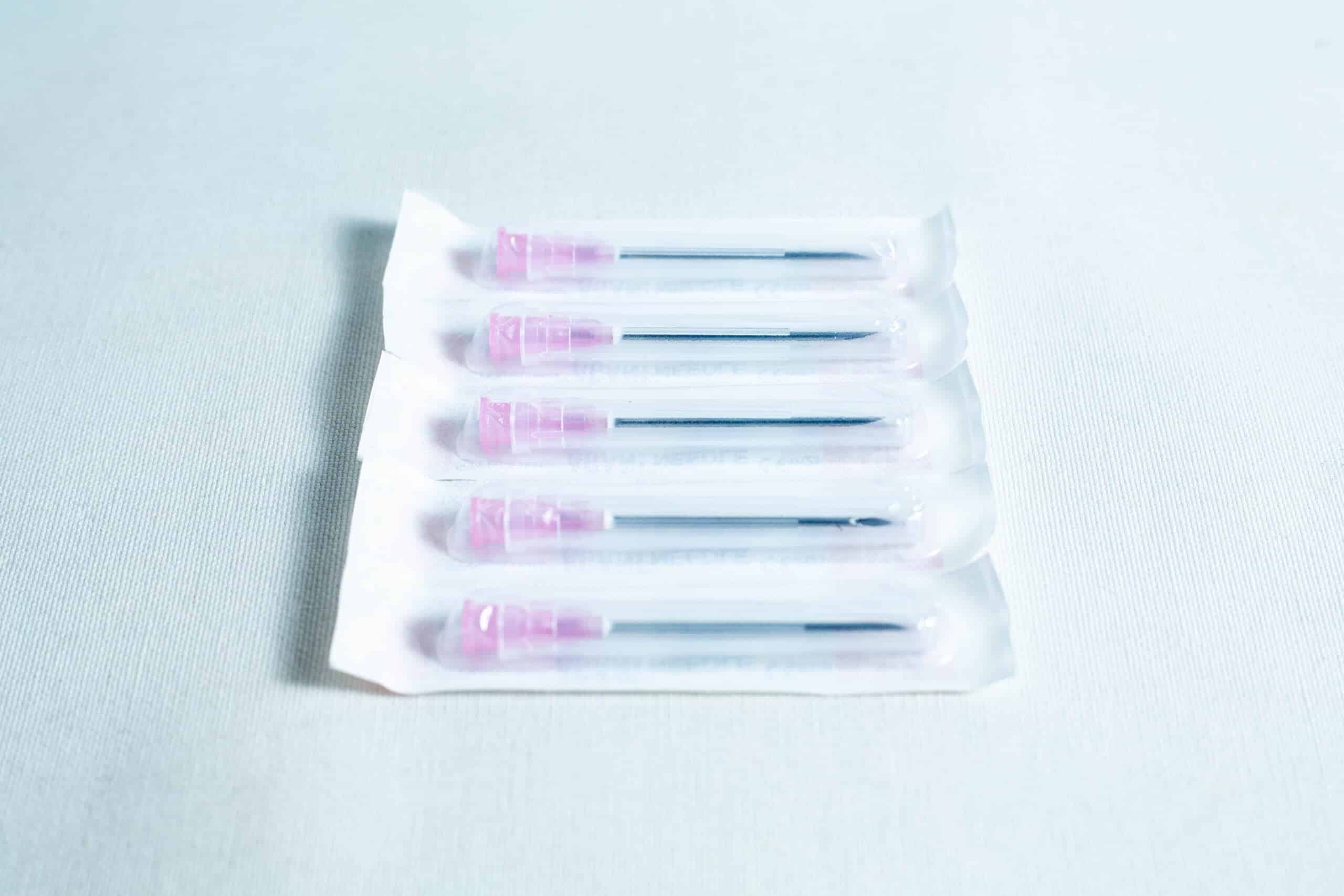 A box of 100 sterile 18G hypodermic needles, each with a pink cap and individually wrapped, is displayed on a white surface.