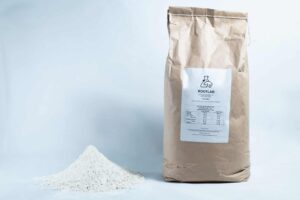 A bag labelled ROOTLAB, containing Brown Rice Flour 5kg Organic Food Grade, stands beside a mound of fine white powder on a white background.