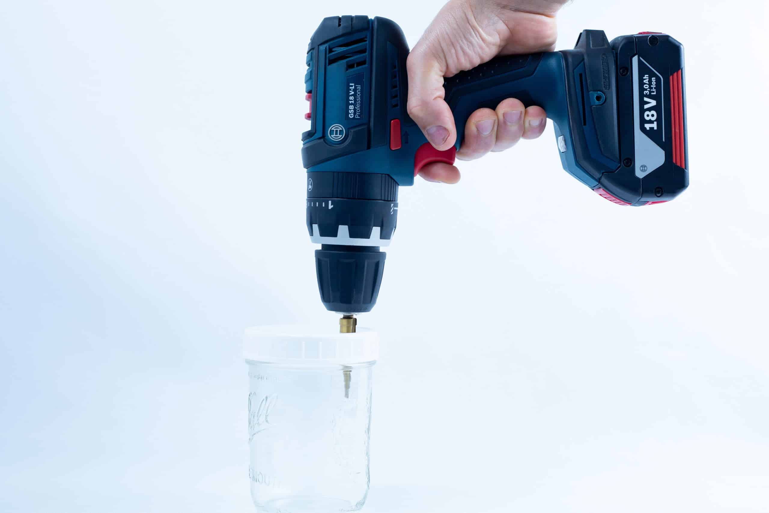A hand holds a Drill Bit for hole sizes 3mm-12mm Titanium Coated attached to a power drill, positioned above a glass jar on a white background.
