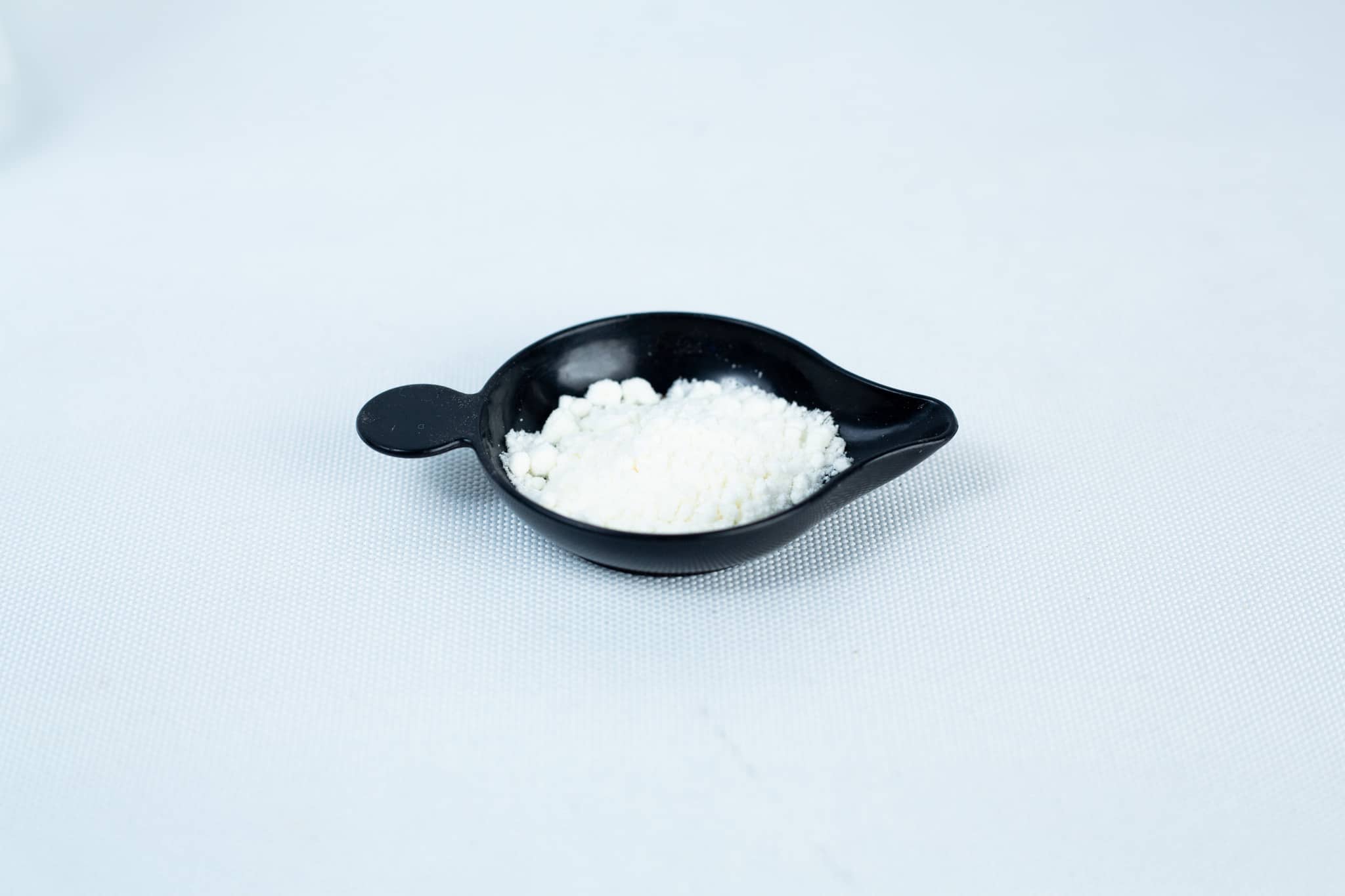 A small black dish holds Antibacterial Powder for mushroom cultivation, seen as a fine white substance on a plain white surface.