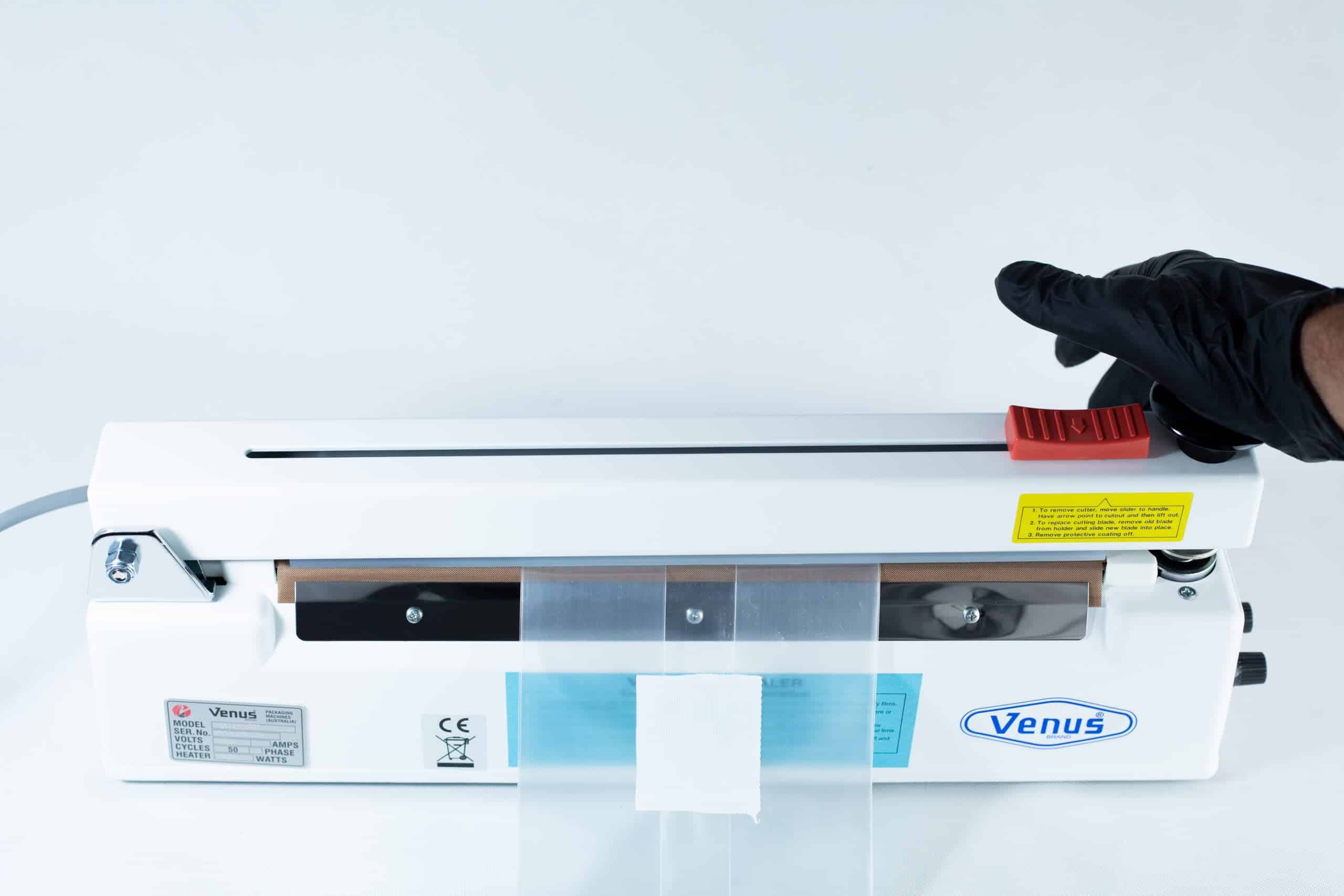 A gloved hand operates a Heat Sealer 30cm Seal Width 5mm with cutter and magnet, sealing a plastic bag securely inside the machine.