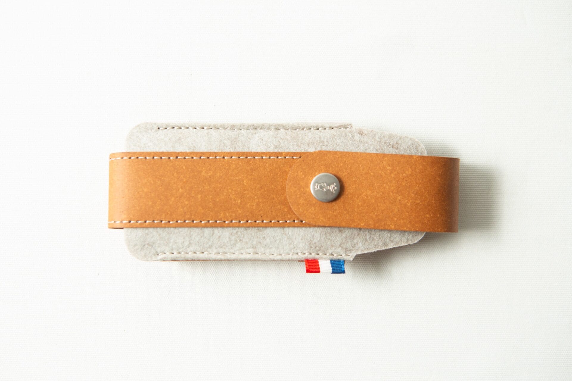 The Opinel Sheath - Brown, made from tan leather and grey felt with a button fastening, rests on a white surface.