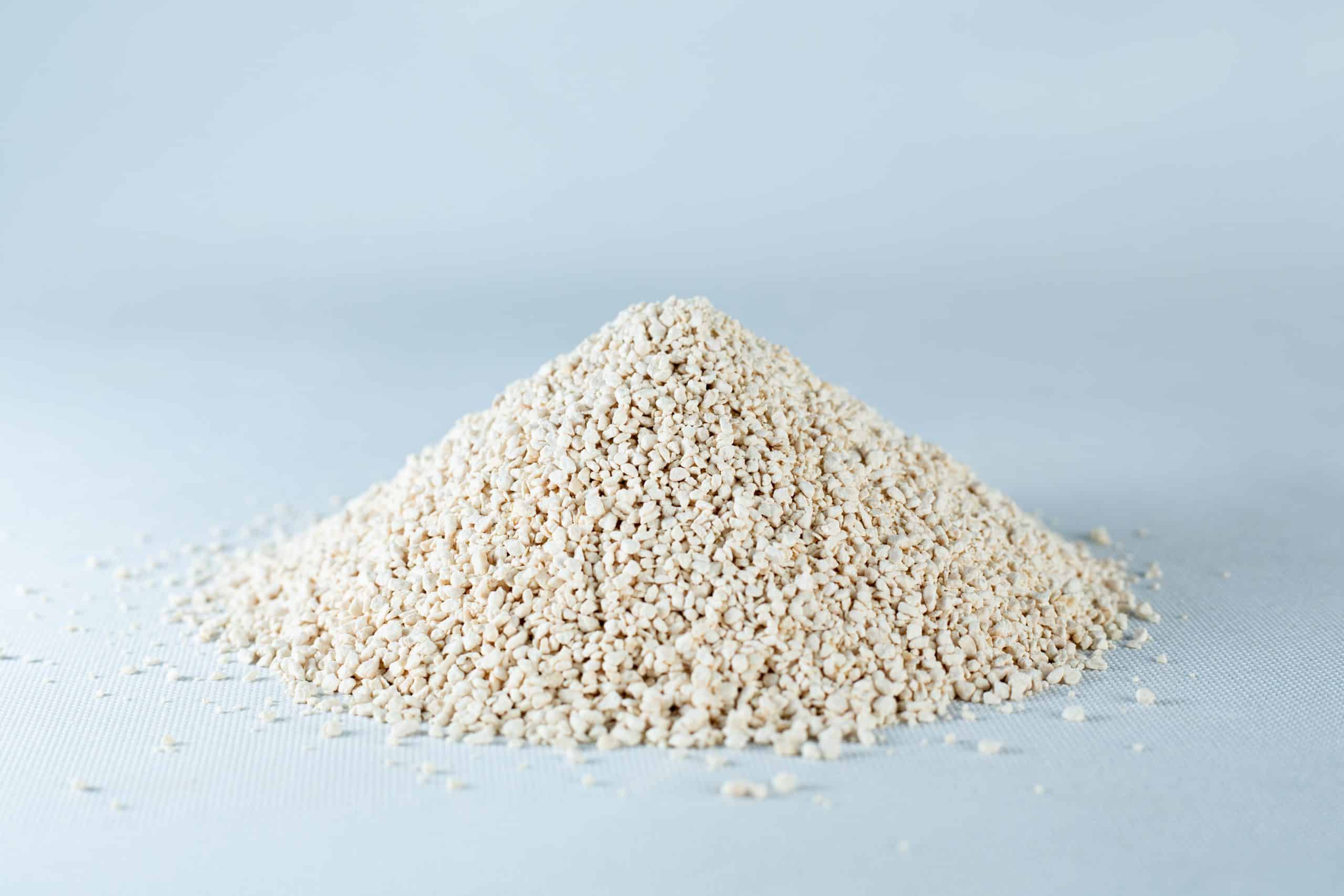 A small mound of Natural Gypsum coarse grade mushroom supplement grain and substrate, featuring beige, granular powder, rests on a light grey surface.
