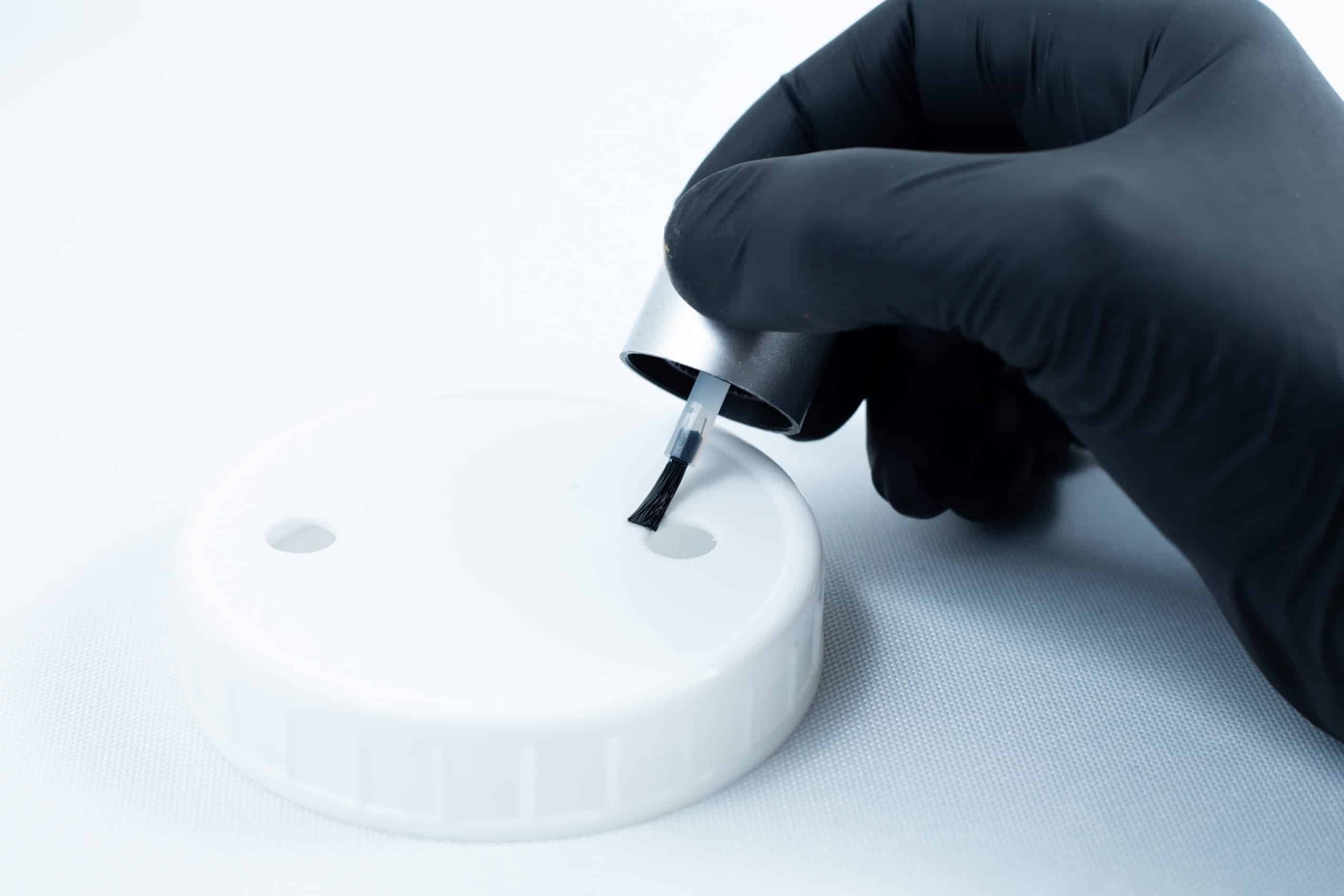 A gloved hand uses a brush to apply Primer 94 multisurface for 3M glue 10ml onto a white plastic lid with two holes, preparing it for adhesive application.