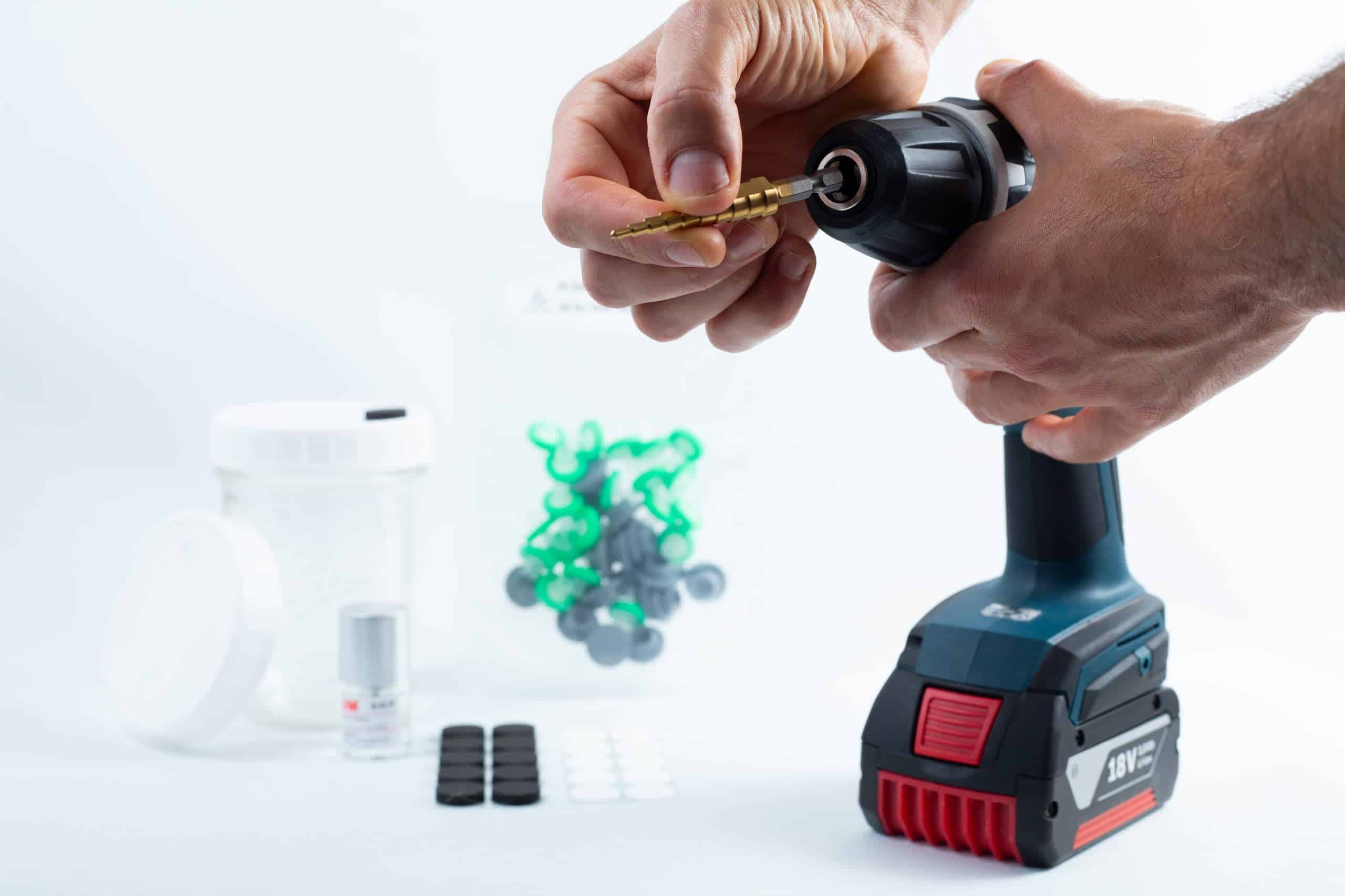 Hands insert the Drill Bit for hole sizes 3mm-12mm Titanium Coated into a cordless drill; various small items are placed on a white surface in the background.