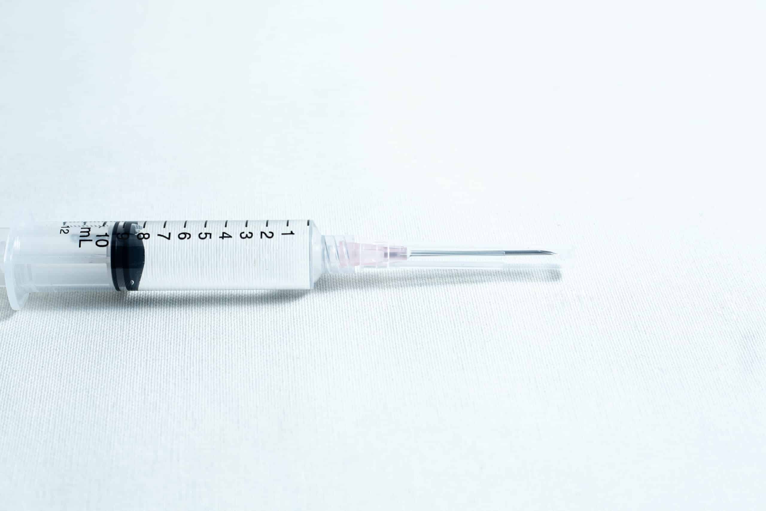 A syringe with clear liquid rests on a white surface, fitted with a sterile 18G hypodermic needle from a box of 100pcs.