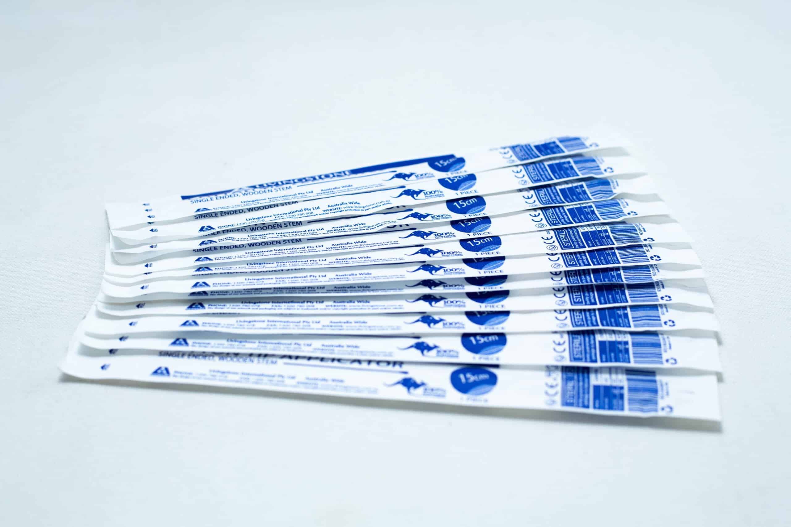 Several individually wrapped sterile cotton bud products in white and blue packaging are arranged on a white surface.
