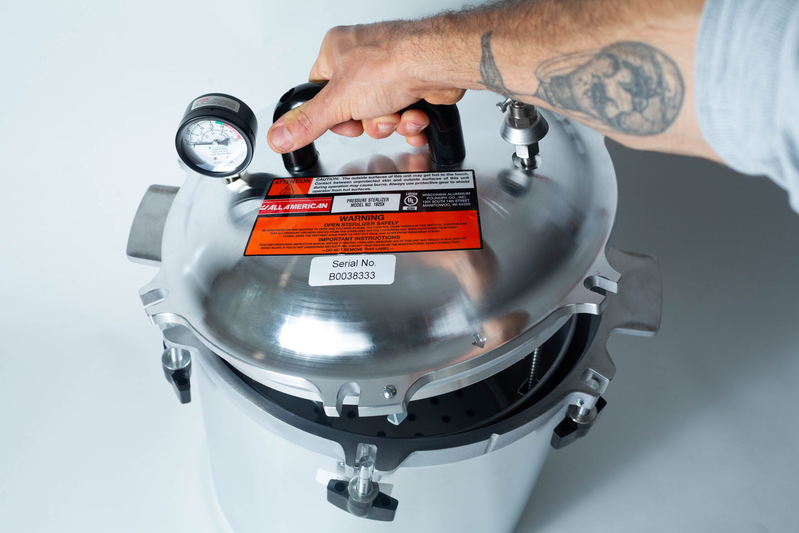 A tattooed hand holding the lid of a stainless steel pressure cooker with a gauge and warning labels.