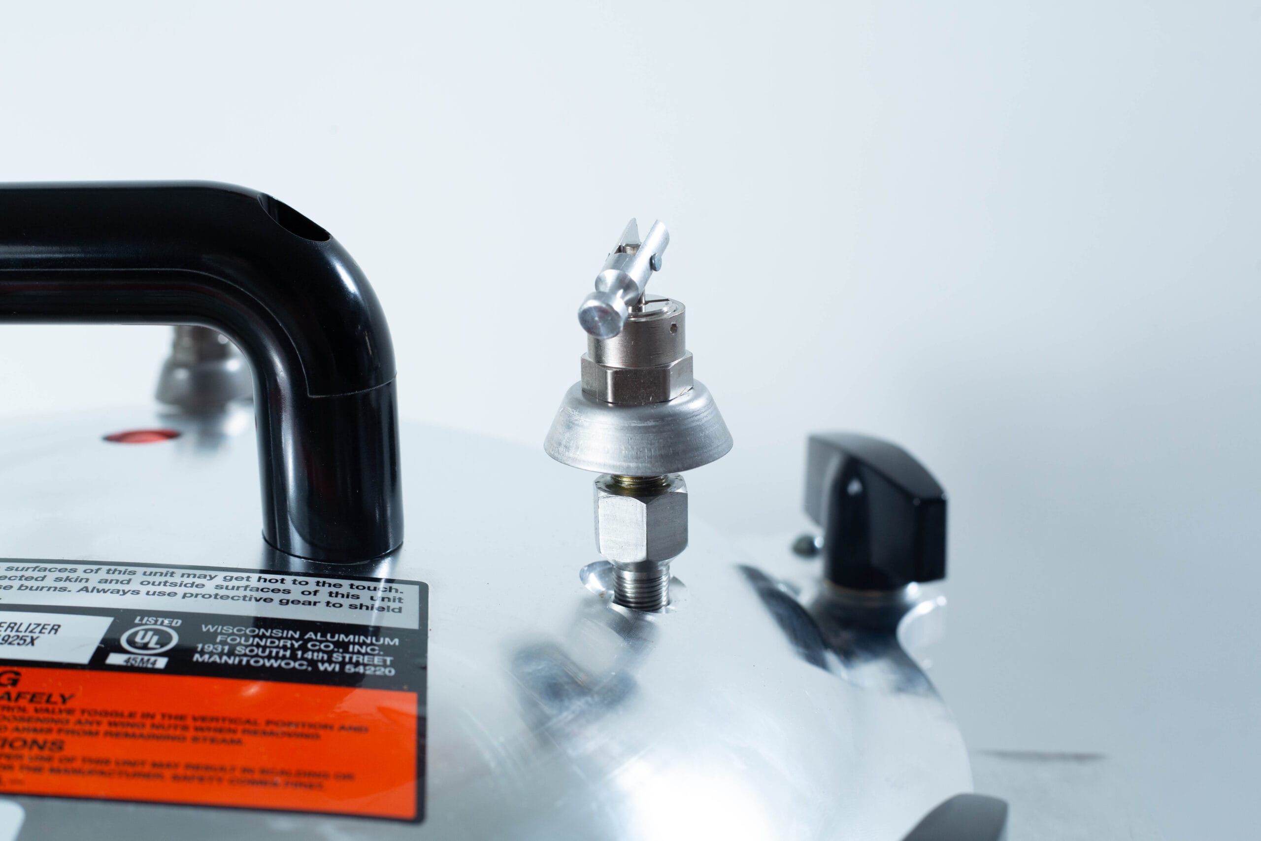 Close-up of an All American Wingnut pressure cooker lid featuring the pressure valve, handle, and safety instructions label.