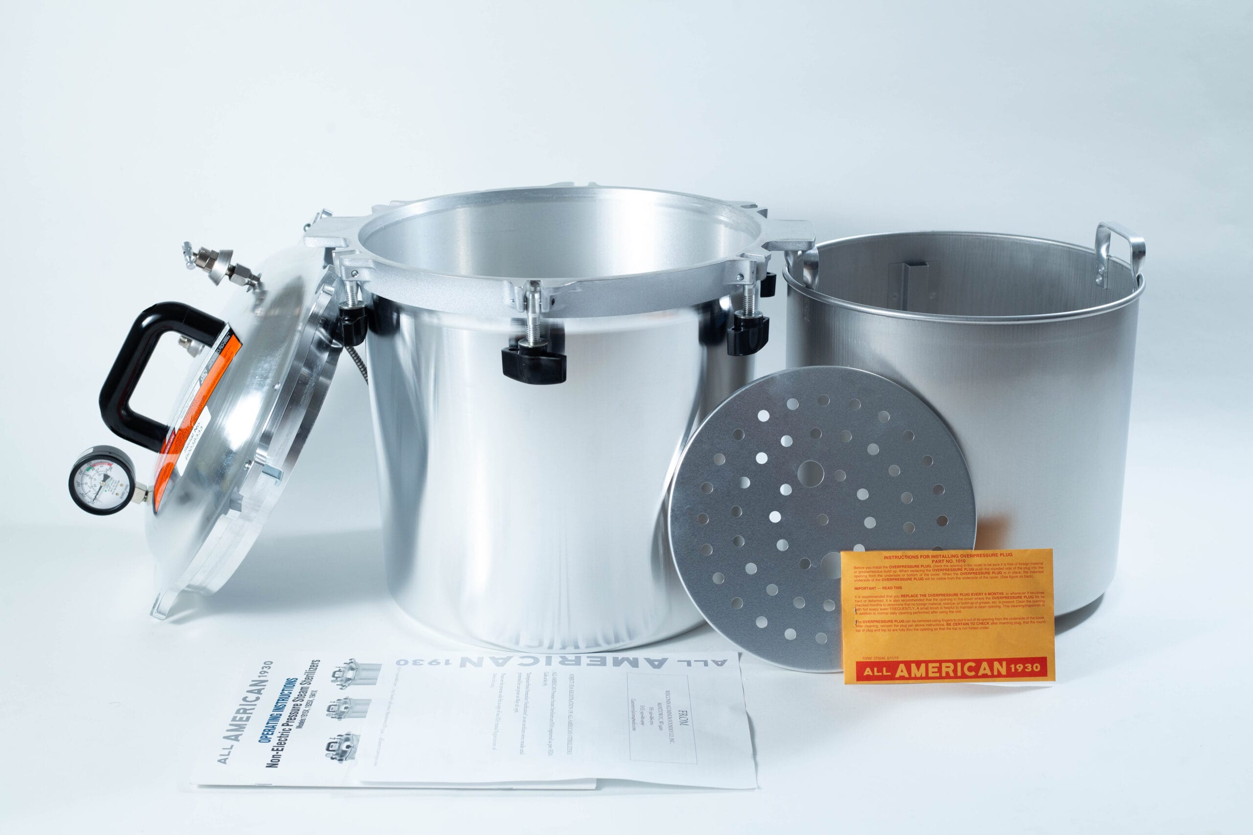 A silver pressure cooker with lid, inner pot, rack, instruction manual, and orange card displayed on a white surface.