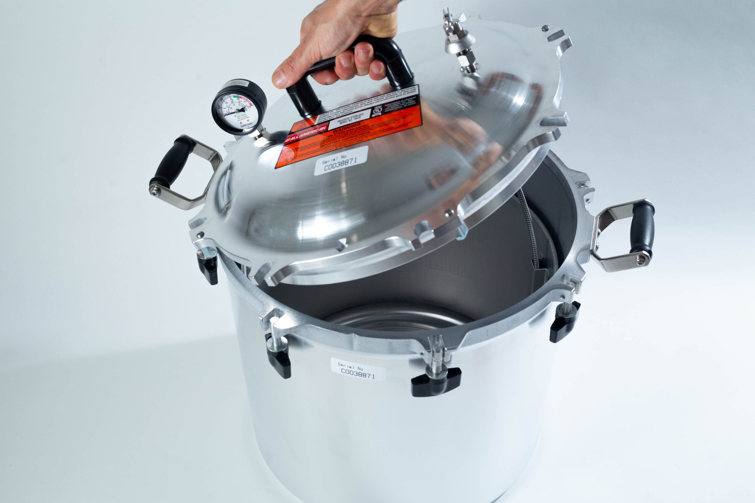 A hand lifts the lid of a large silver pressure cooker with a gauge and black handles.