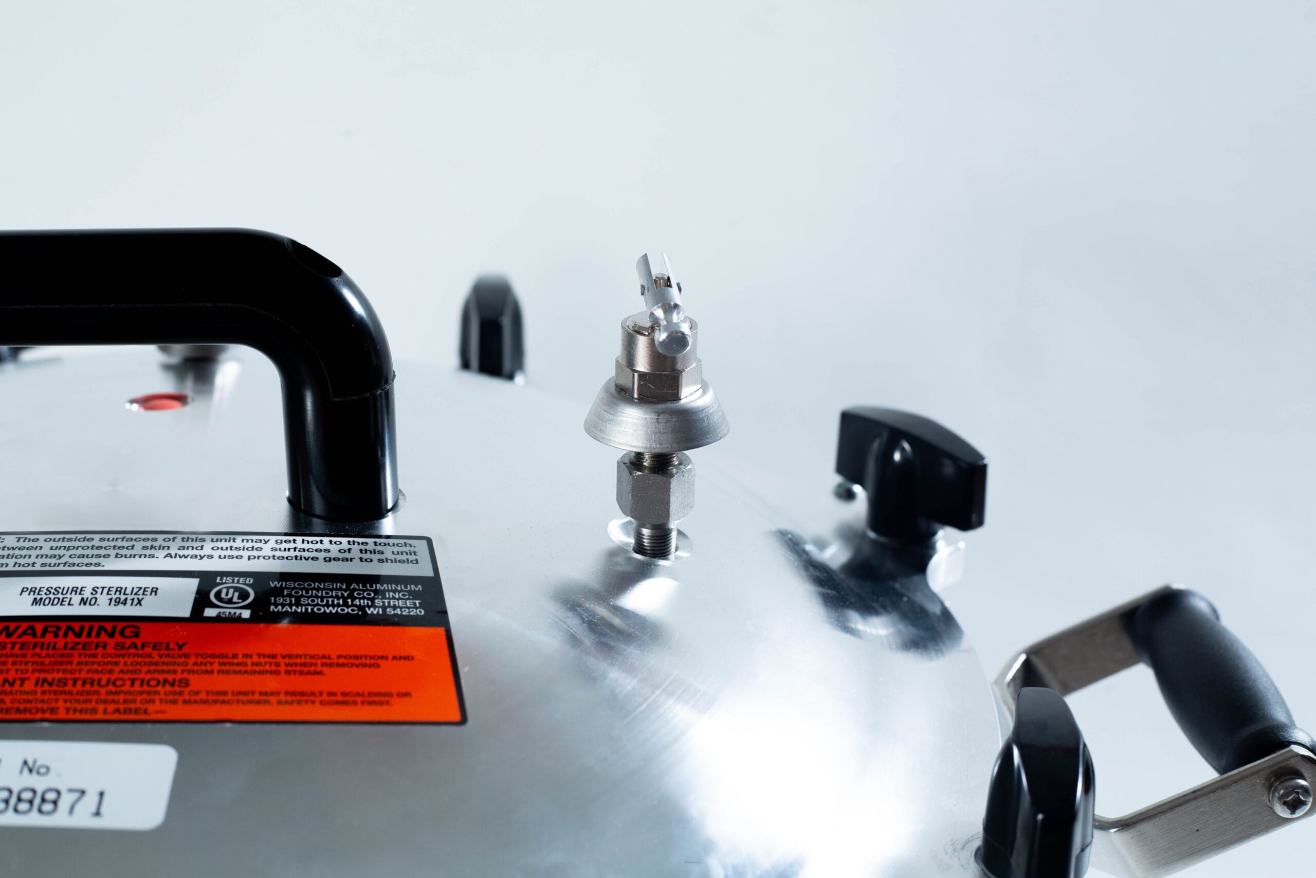Close-up of a metallic pressure cooker lid with handles, safety valve, and warning label visible.