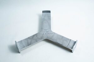 Grey metal object with three prongs and “THIS SIDE UP” engraved at the centre, placed on a white surface.