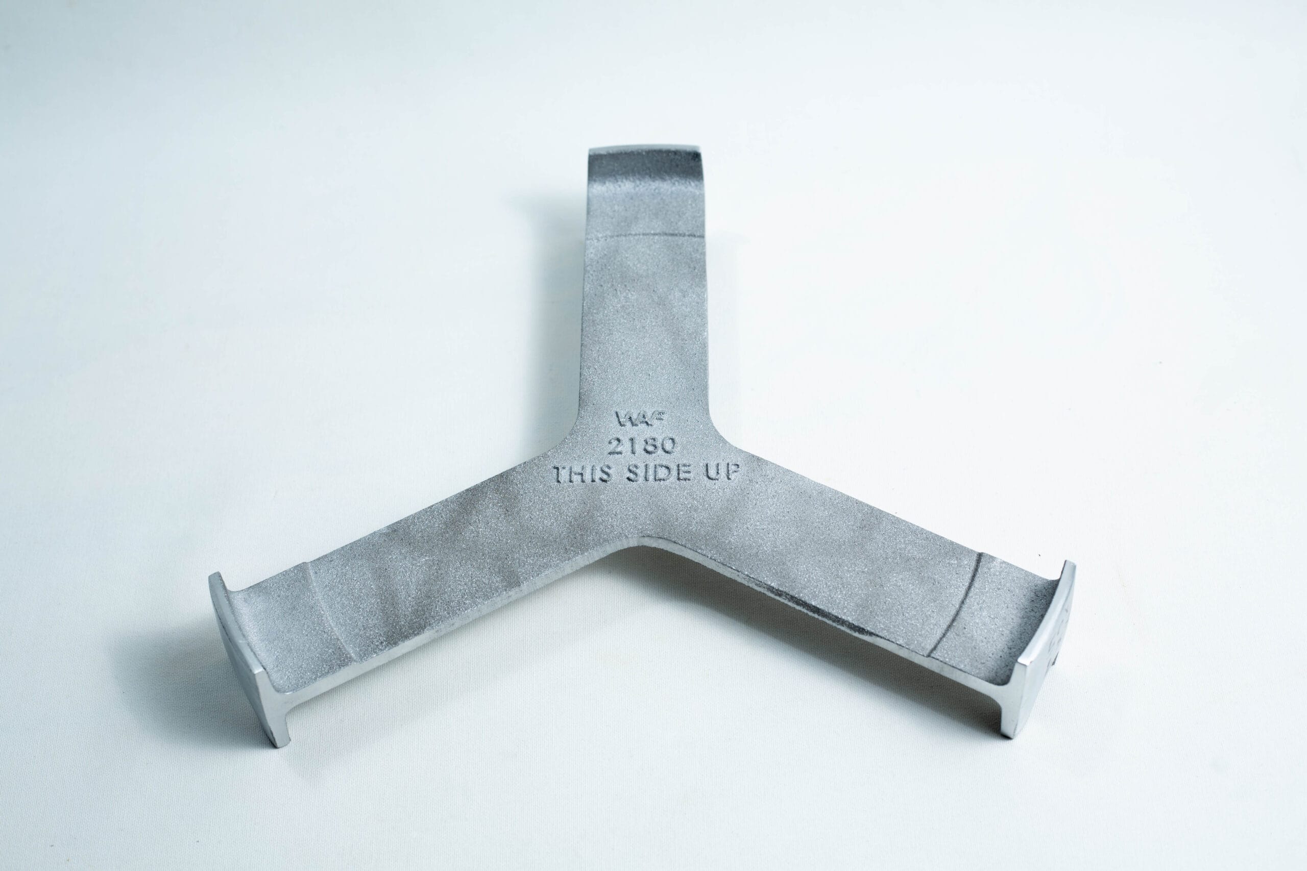 Grey metal object with three prongs and “THIS SIDE UP” engraved at the centre, placed on a white surface.