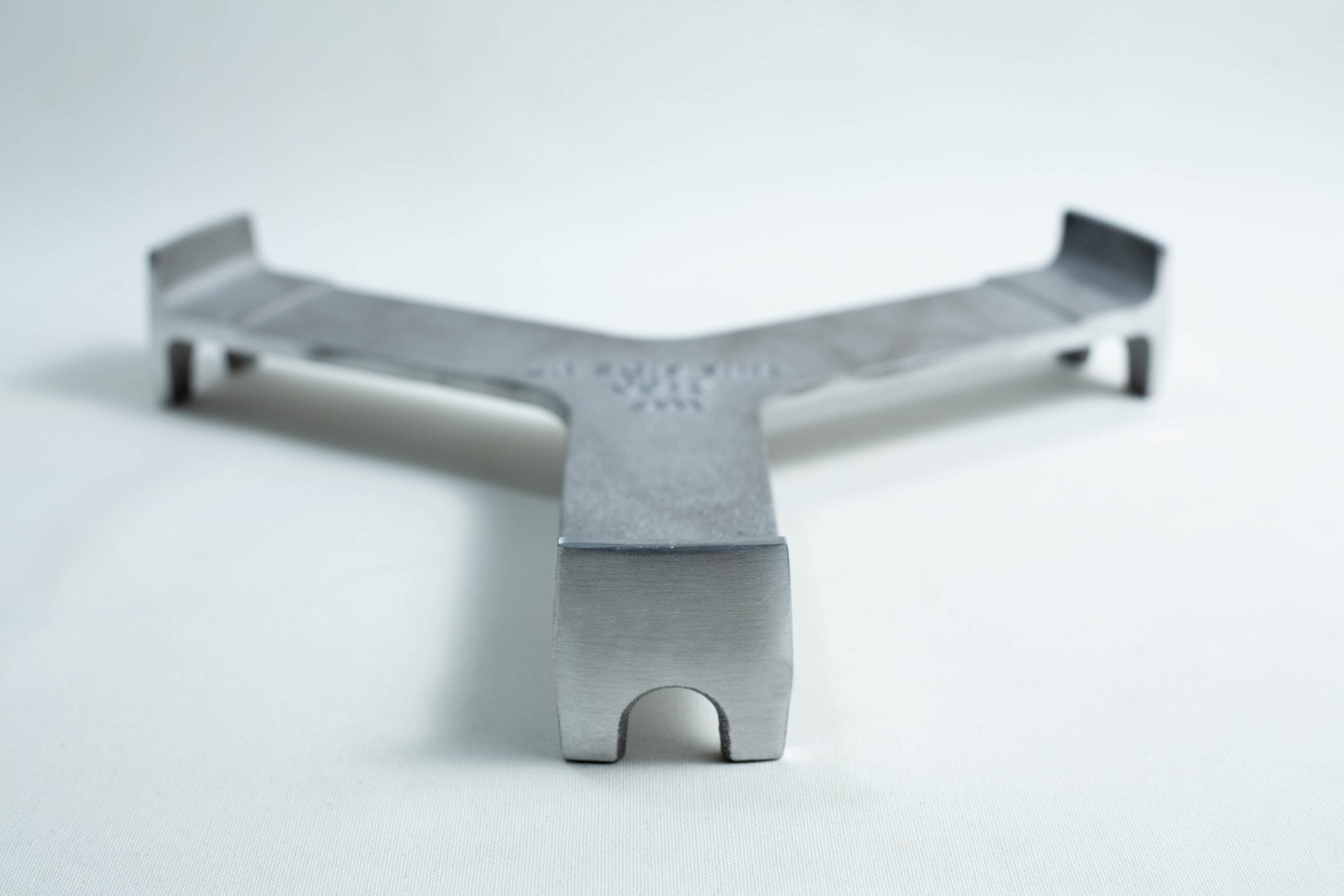 A metal trivet with three arms rests on a white surface, viewed from a low front angle.