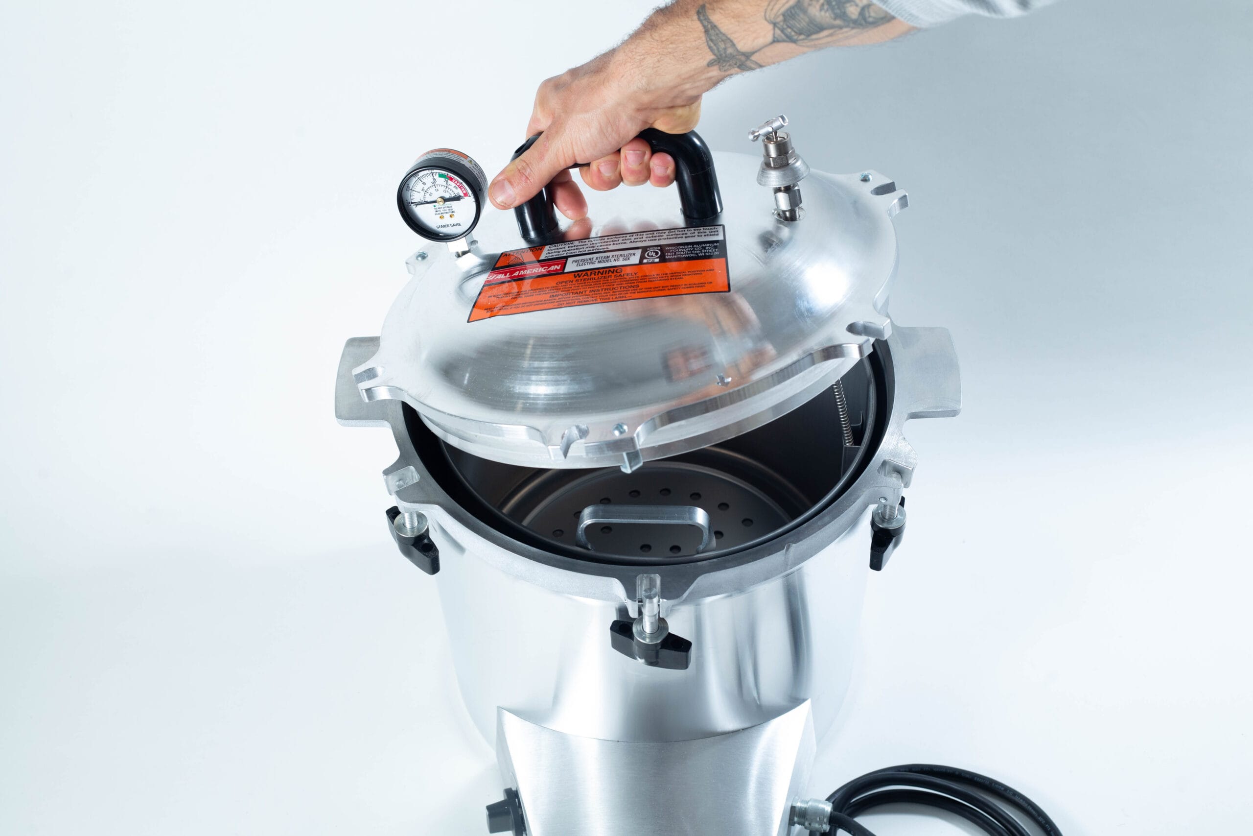 A hand lifts the lid of a stainless steel pressure cooker with a gauge and safety label on top.
