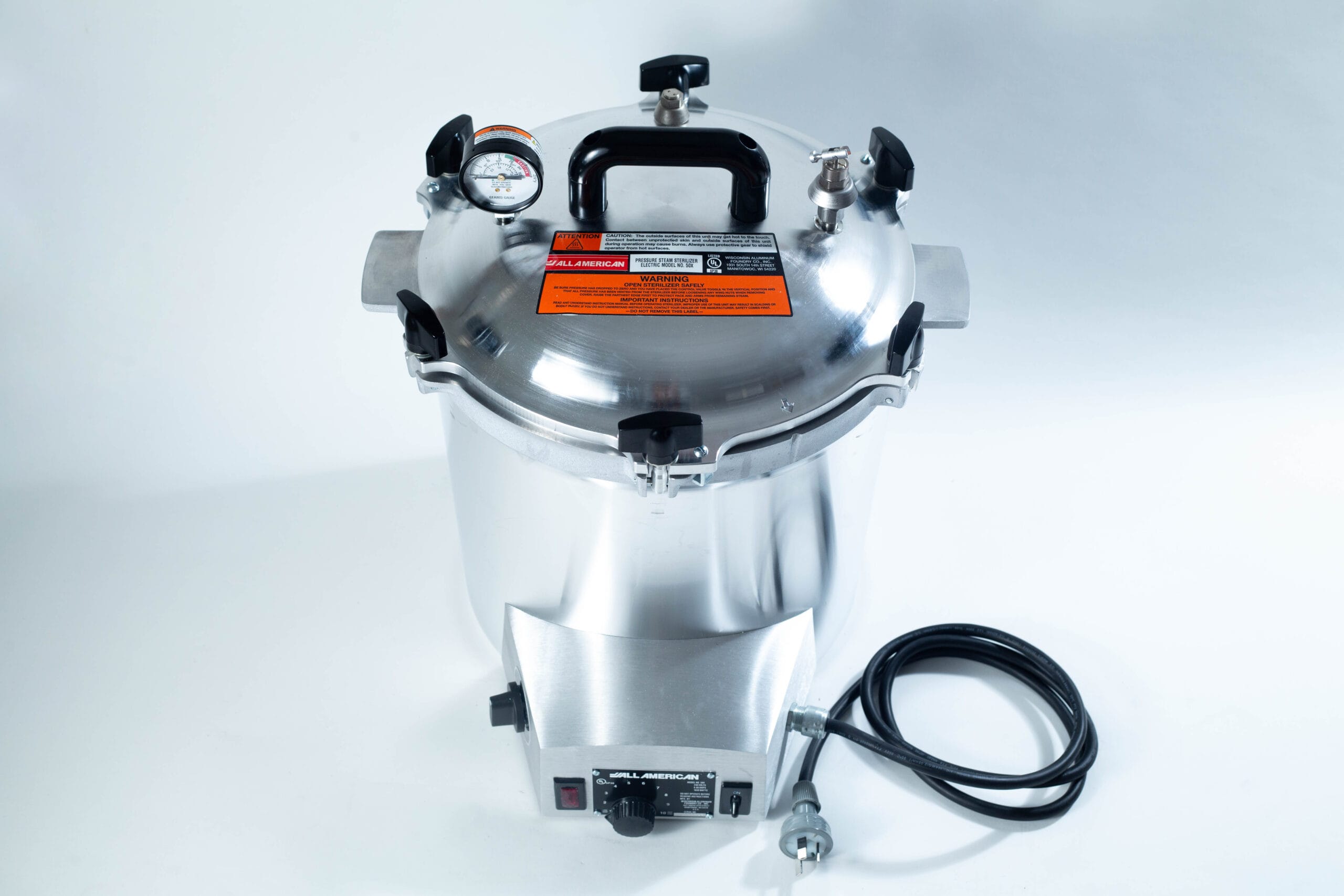 A stainless steel pressure cooker with a gauge, black handles, and an attached power lead on a white background.
