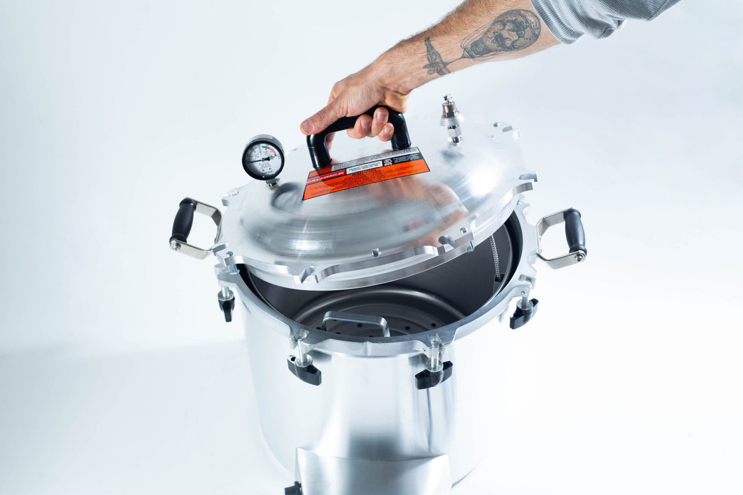 A hand lifts the lid of a stainless steel pressure cooker with a gauge on top.