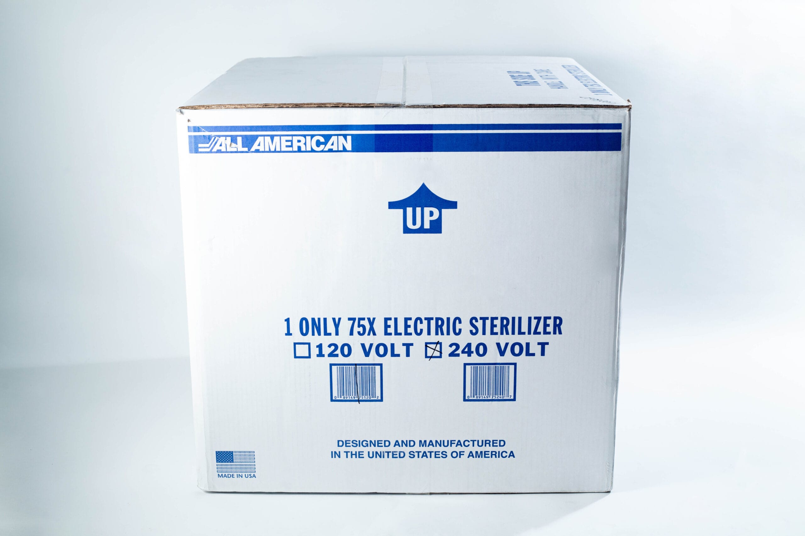 A cardboard box labelled All American containing a 75X electric steriliser, marked UP with voltage options.
