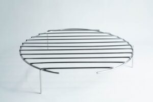 Round metal grill rack with three legs, standing on a white surface, photographed against a plain background.