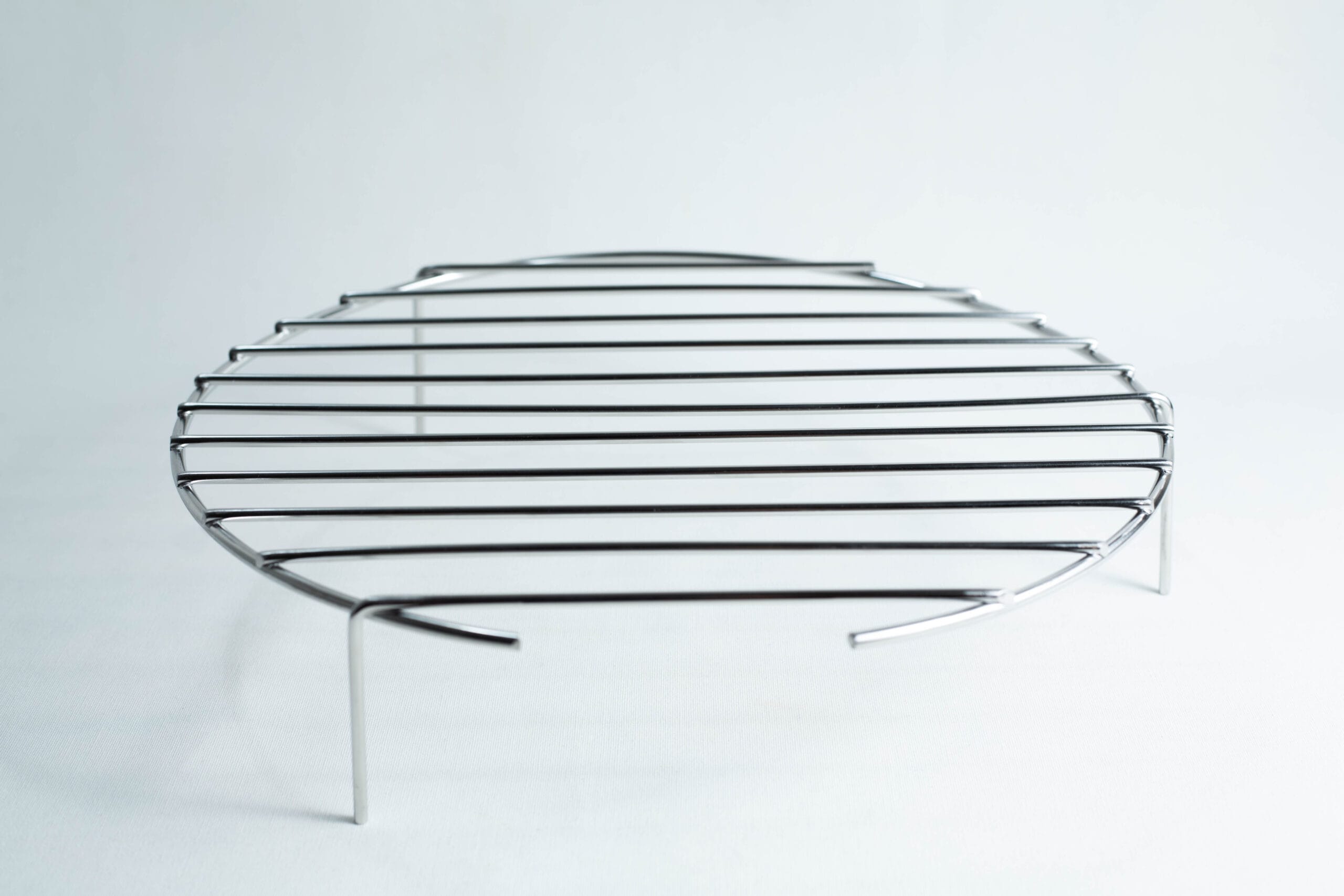 Round metal grill rack with three legs, standing on a white surface, photographed against a plain background.
