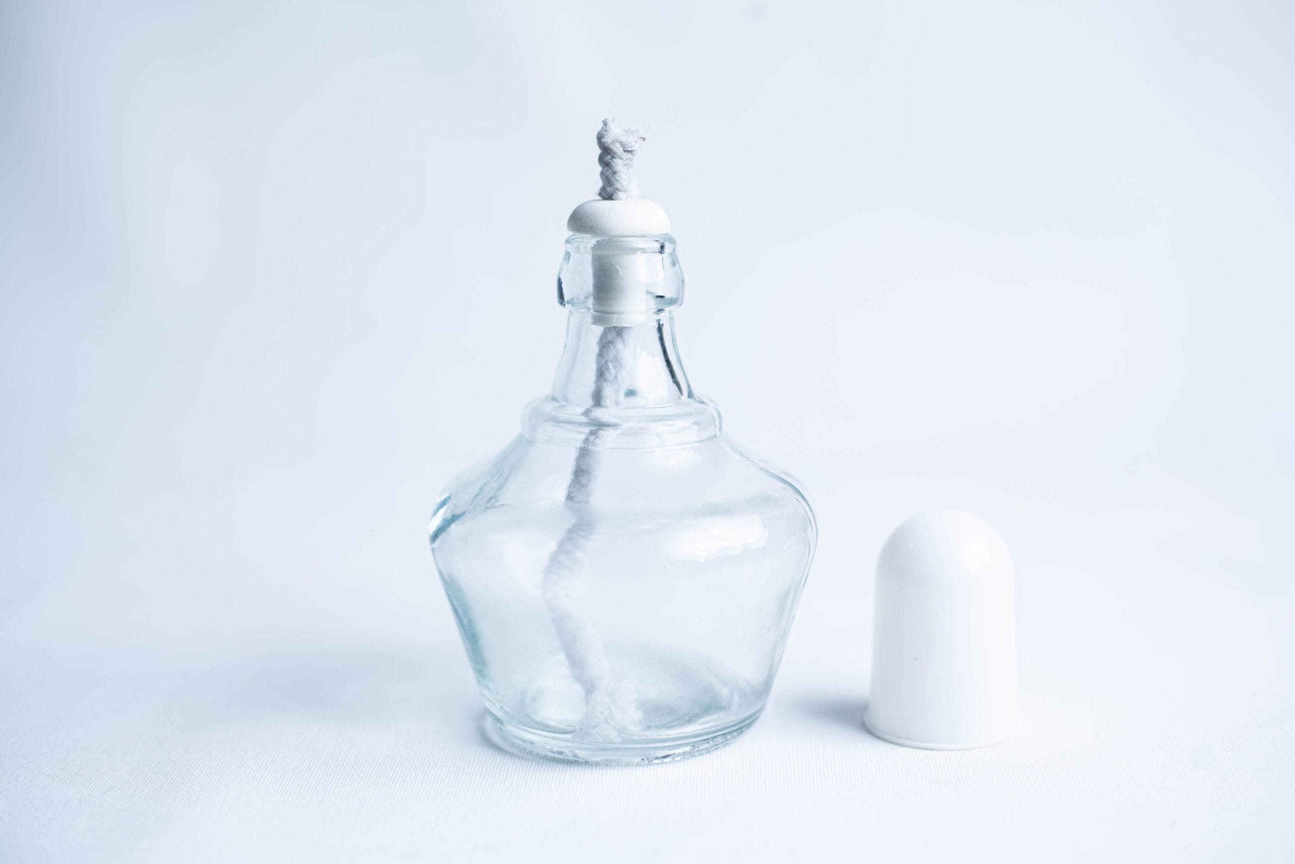 Clear glass perfume bottle with a stopper and white cap placed beside it on a white background.