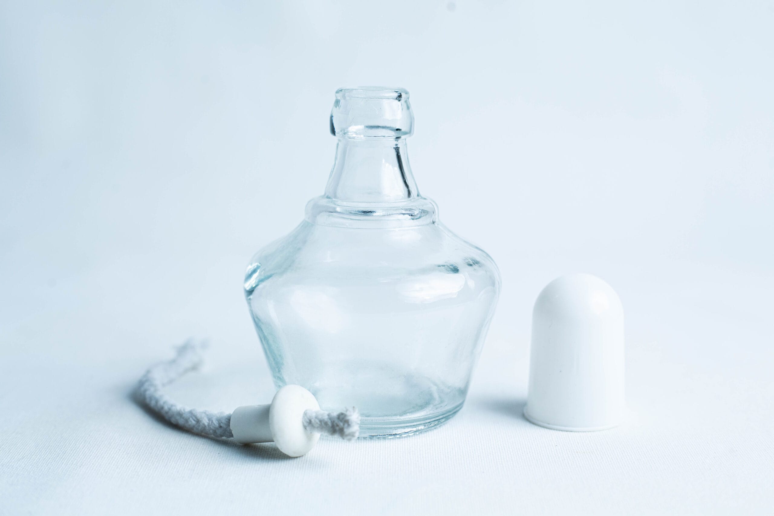 A small empty glass bottle with its white cap and a wick placed beside it on a white background.