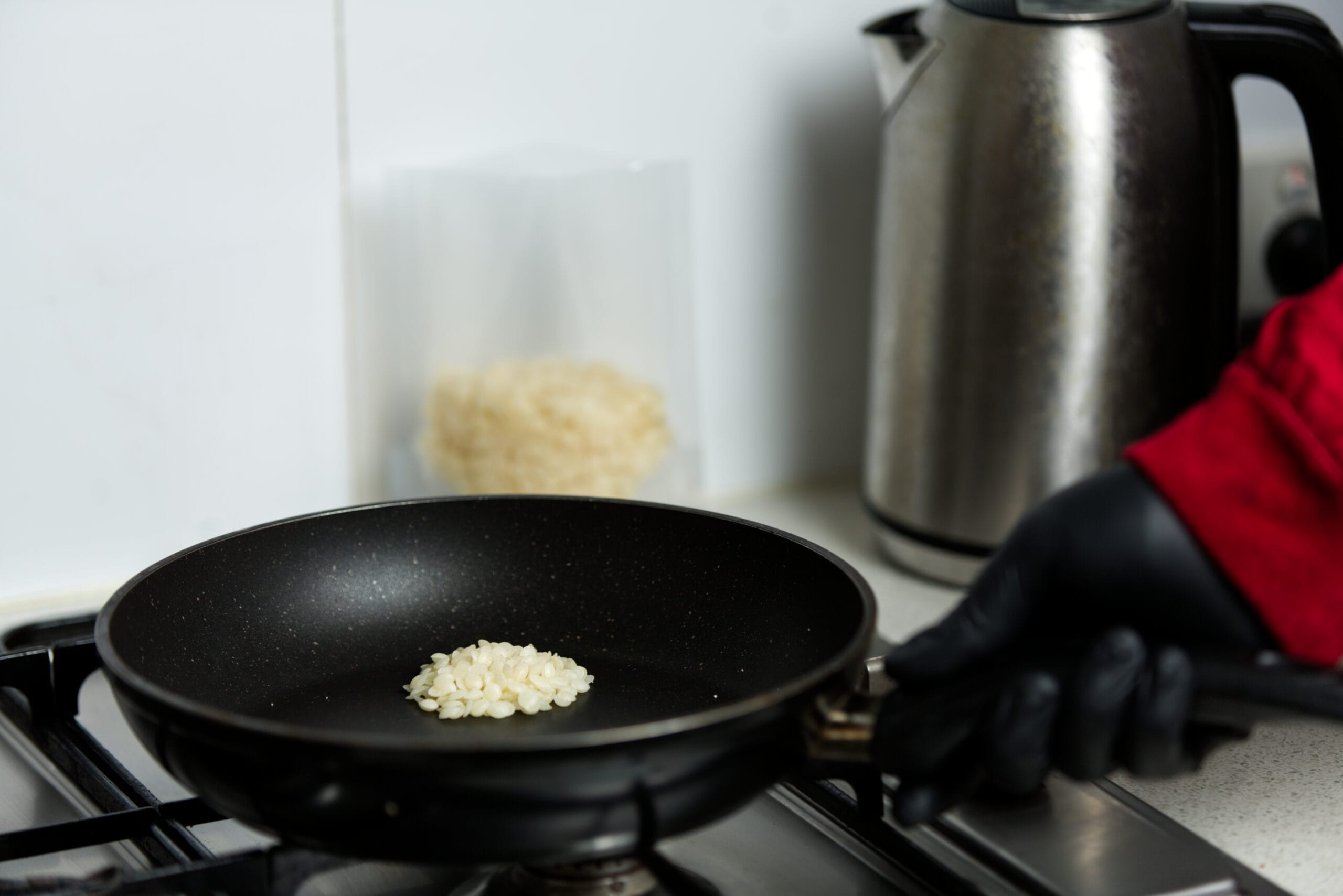 A hand in a black glove holds a pan with rice cooking on a hob next to a metal electric kettle.