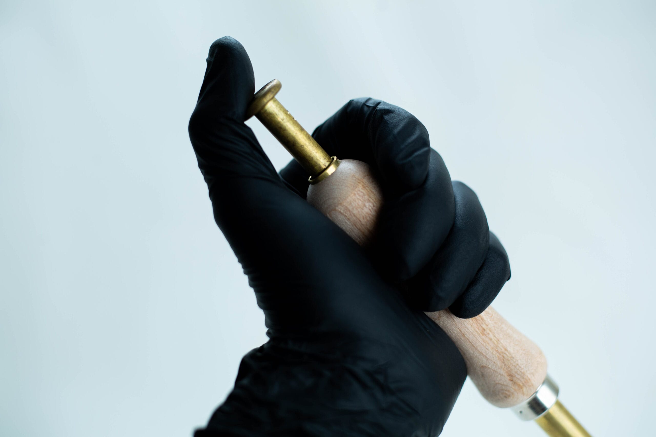 A hand in a black glove holding a brass and wooden tool against a plain light background.