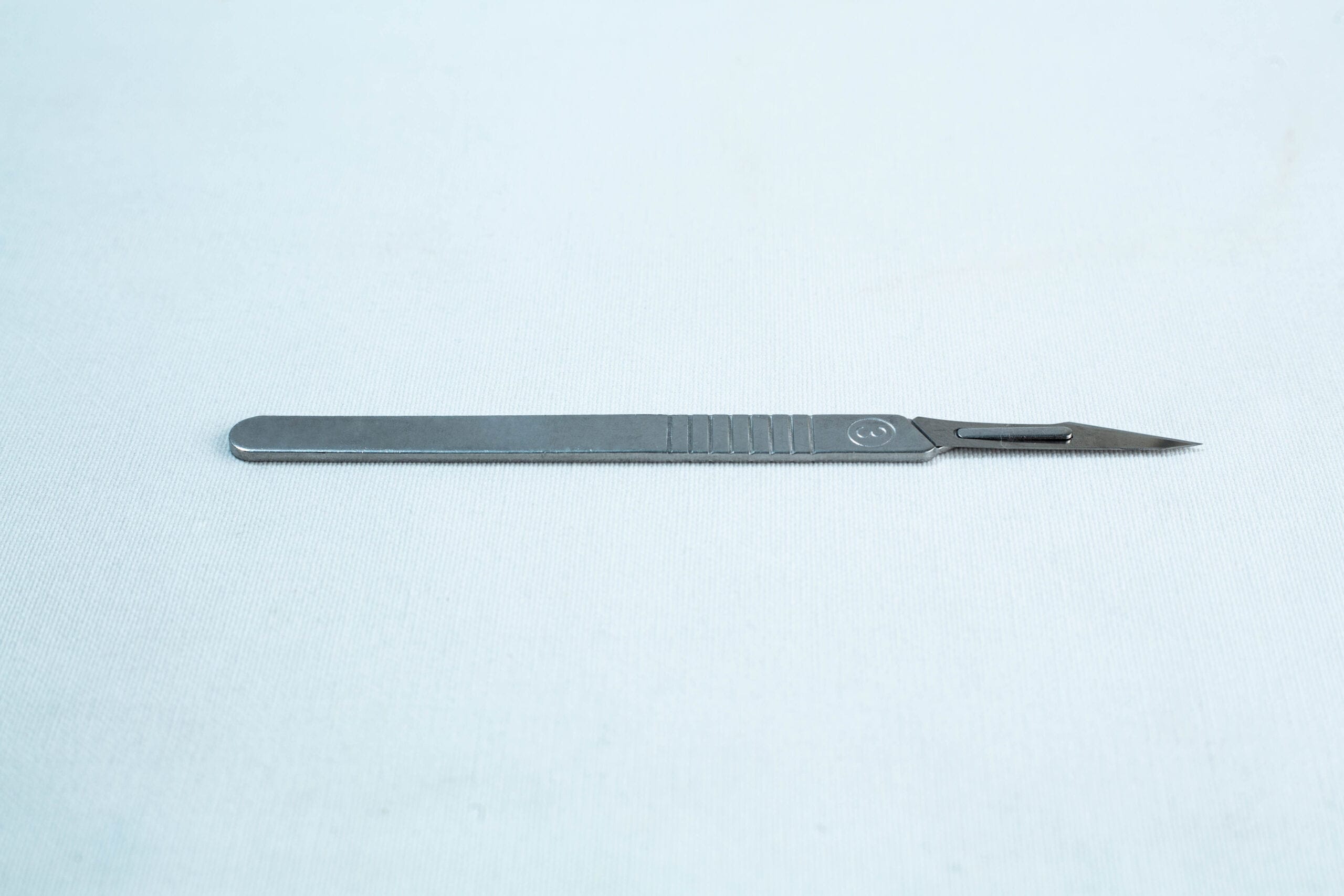 A metal surgical scalpel lies on a plain light surface with its blade facing right.