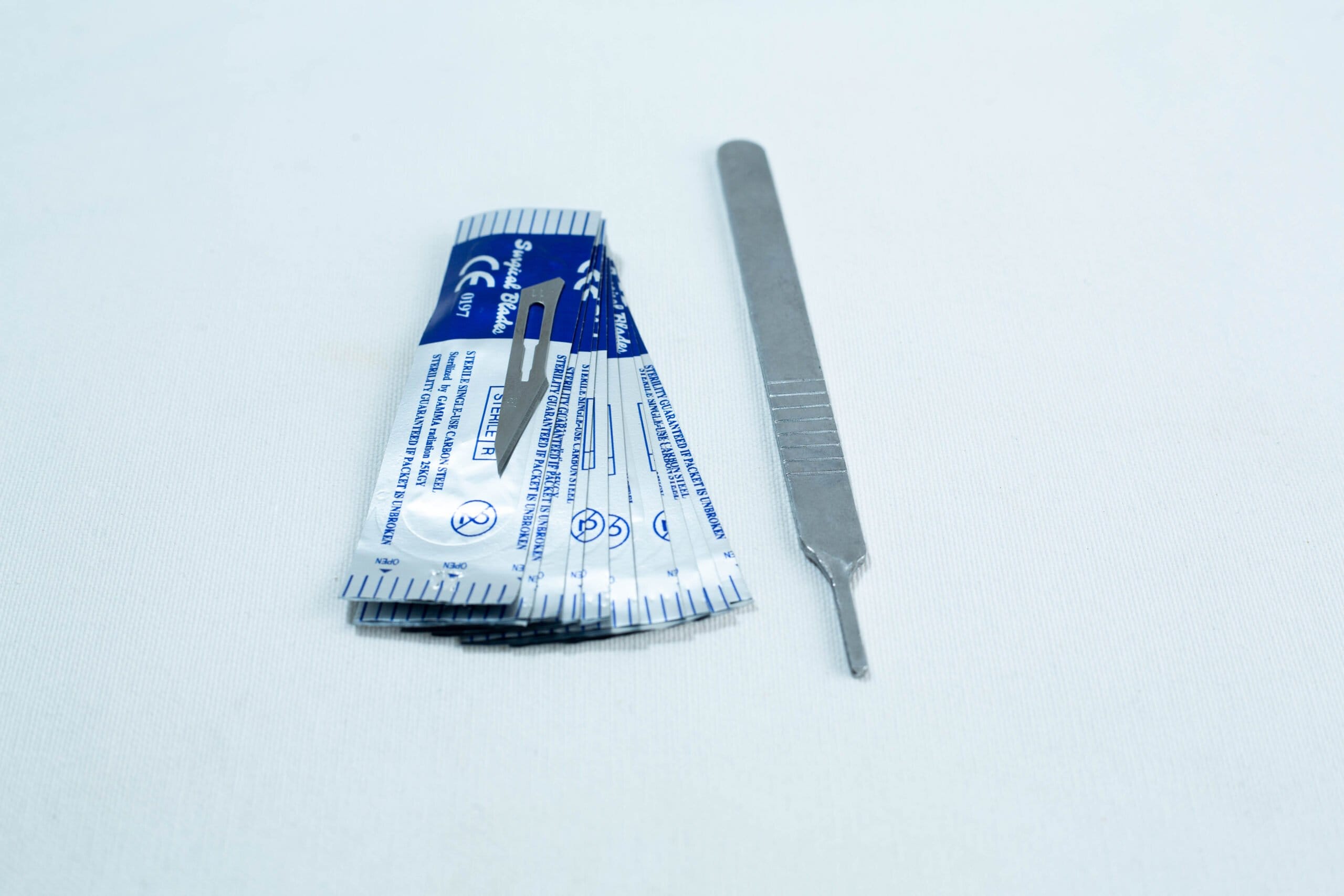 A scalpel handle next to several individually wrapped scalpel blades on a white surface.