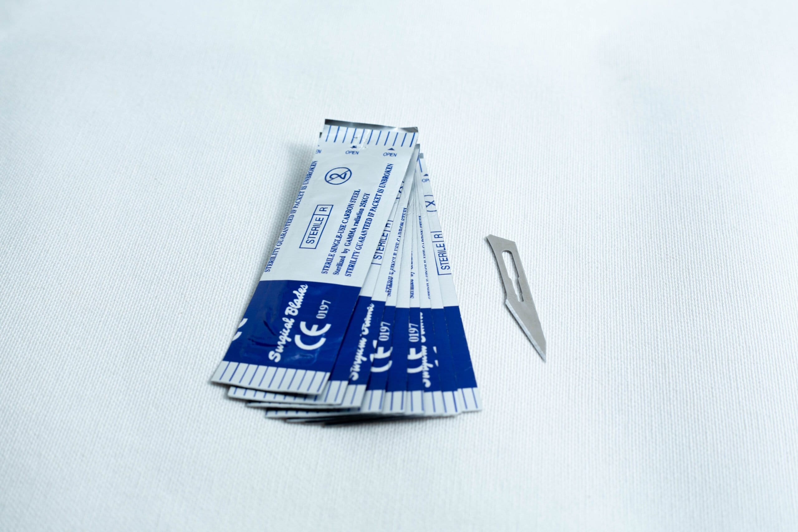 Several wrapped surgical blades and one unwrapped blade on a white surface.