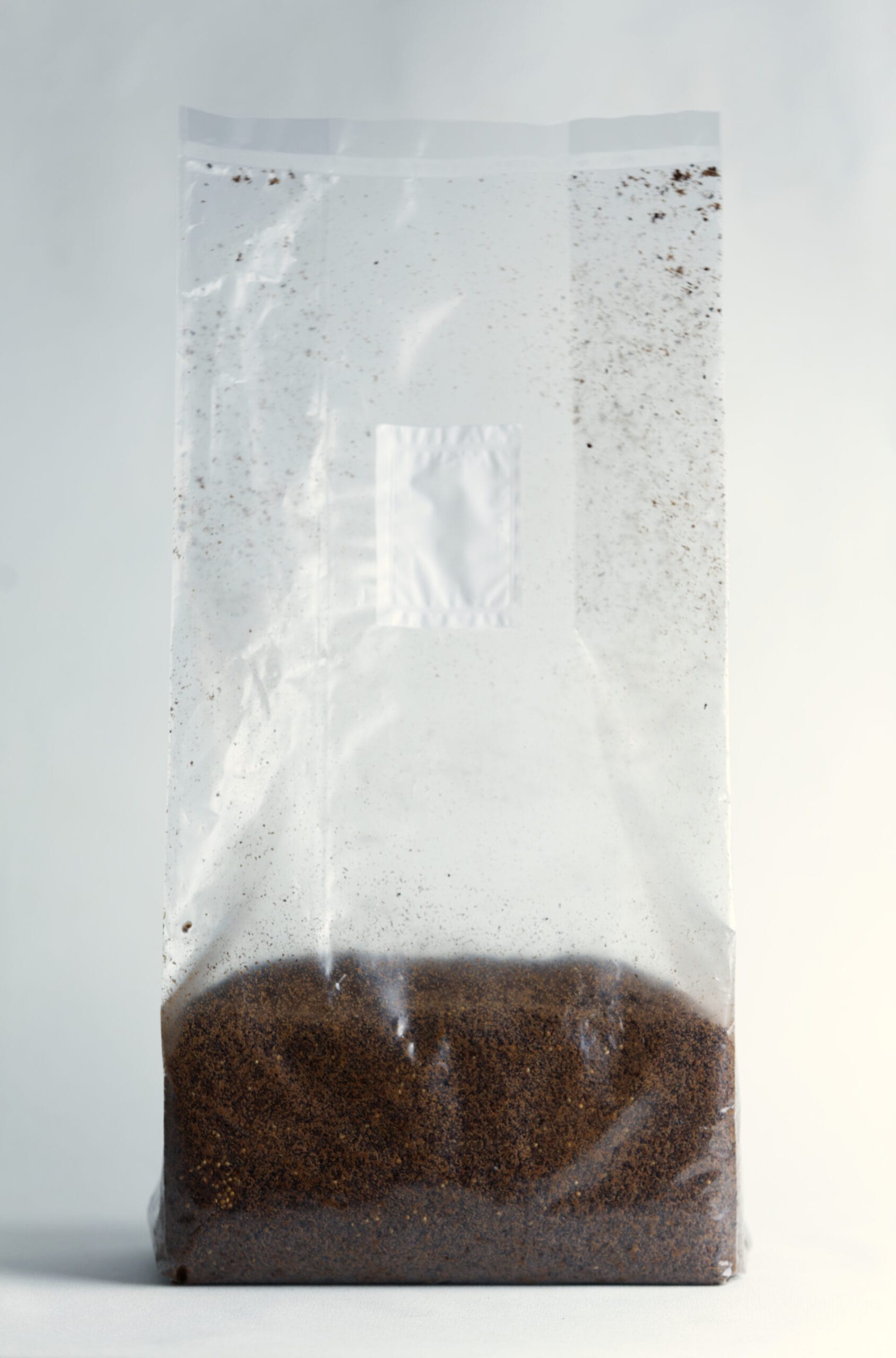 Clear plastic bag filled with brown coffee grounds against a plain background.
