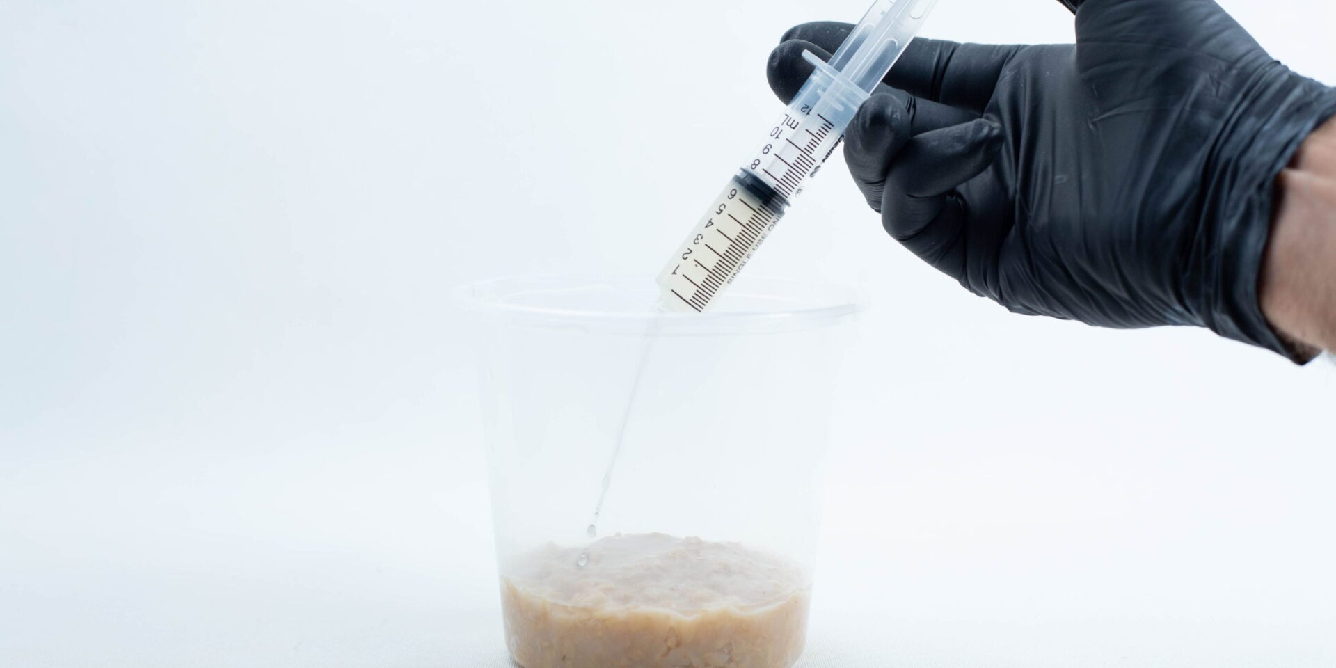 A gloved hand holds a syringe above a plastic container with a beige, lumpy liquid inside.