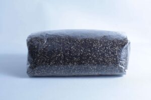 A block of dark soil or compost, perfect for the Monotub Mushroom Growing Kit-Premium, is wrapped in clear plastic and set against a plain light background.
