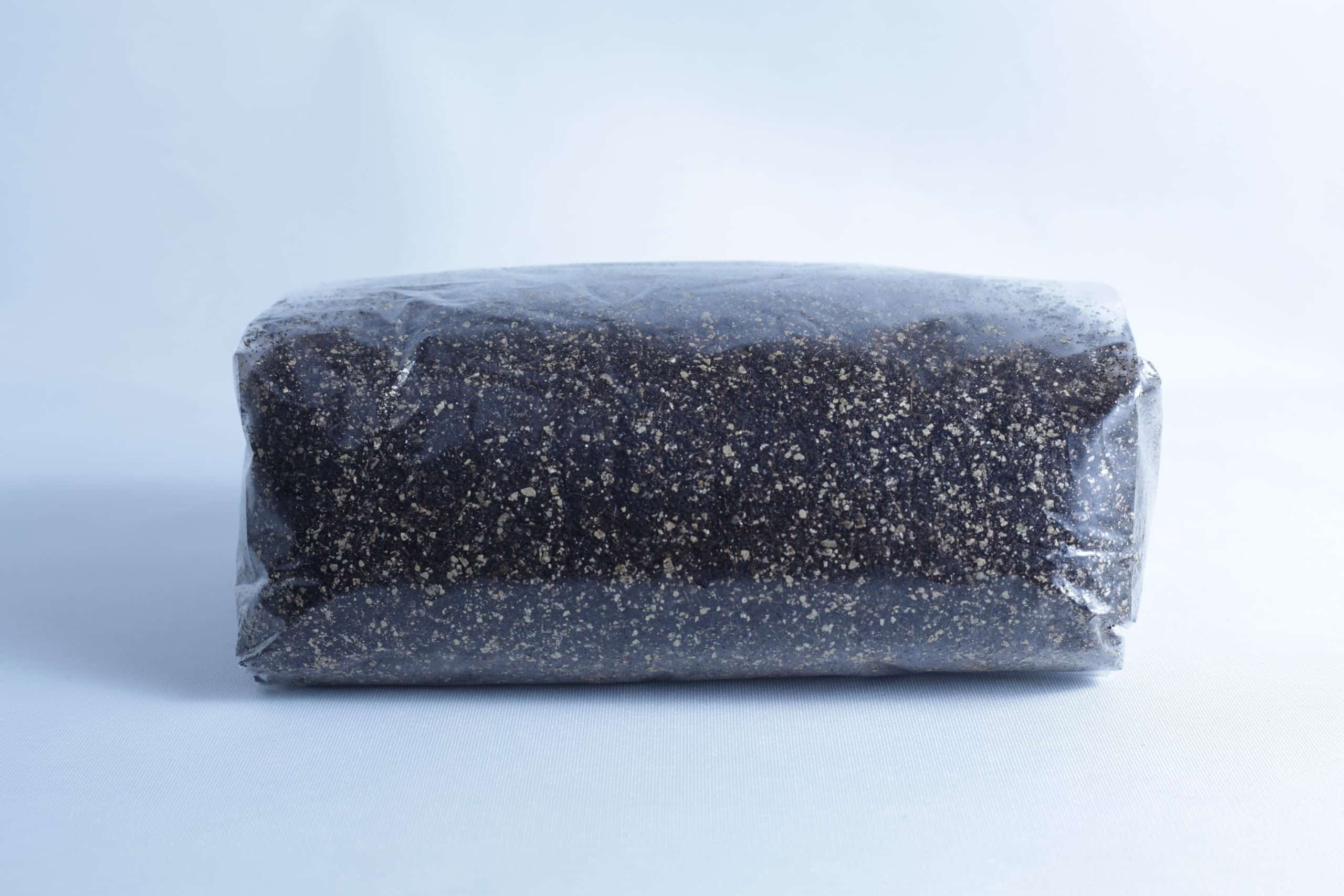 A block of dark soil or compost, perfect for the Monotub Mushroom Growing Kit-Premium, is wrapped in clear plastic and set against a plain light background.