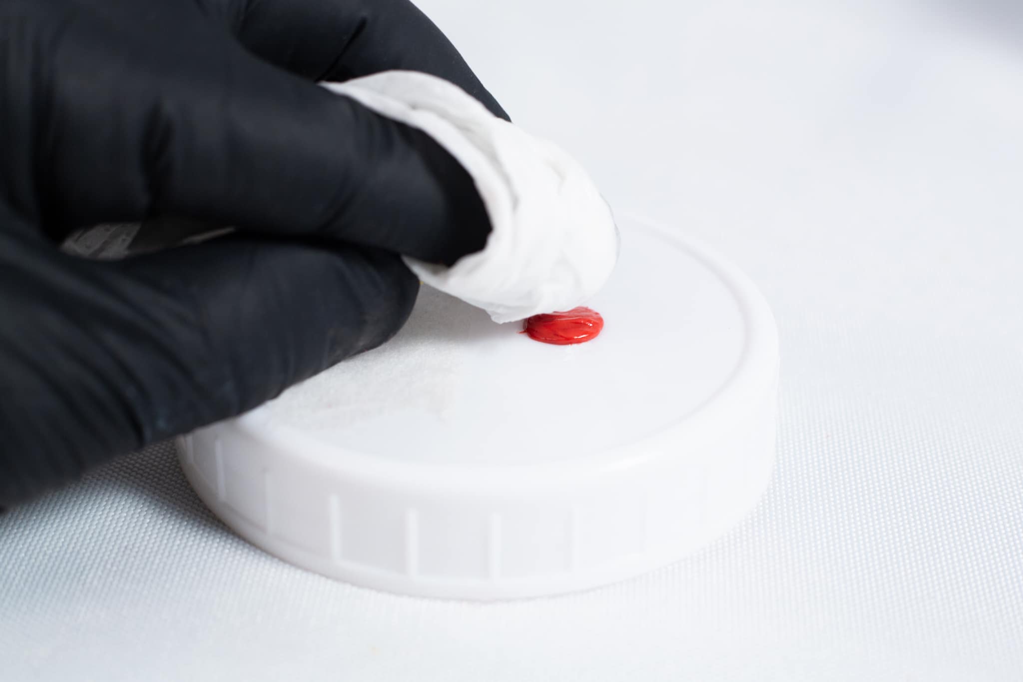 A gloved hand wipes a red stain from a white plastic lid with a tissue, ensuring the surface is clean before applying High Temperature RTV Silicone-Red.