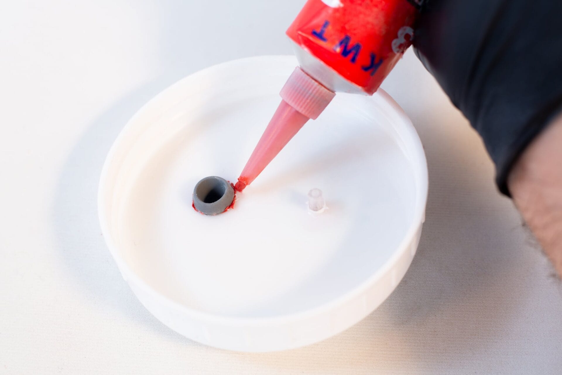 A person dispenses High Temperature RTV Silicone-Red through a nozzle into a small hole of a white circular container, preparing it for sealing.
