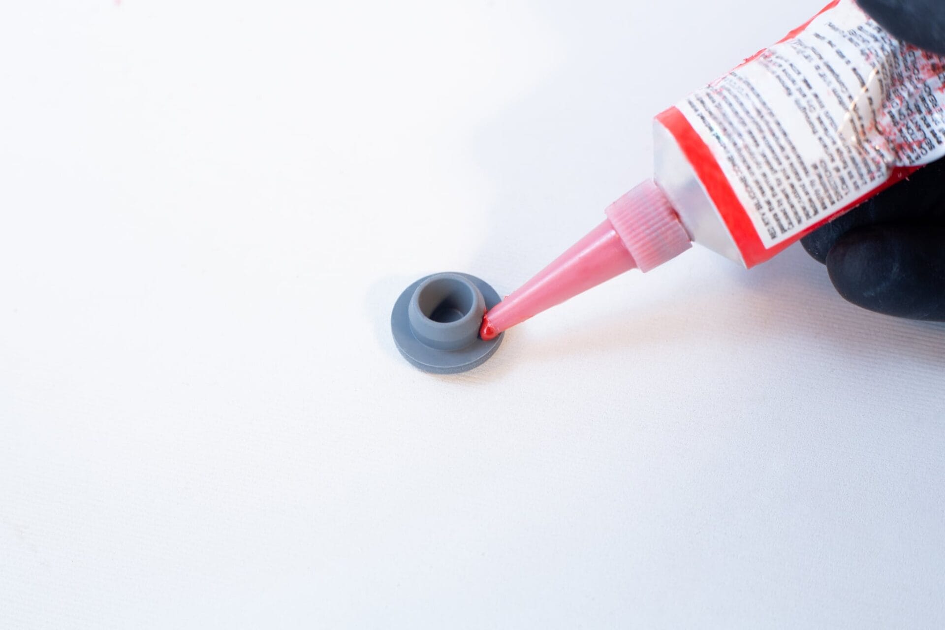 A hand applies High Temperature RTV Silicone-Red from a tube onto a small grey plastic part placed on a white surface.