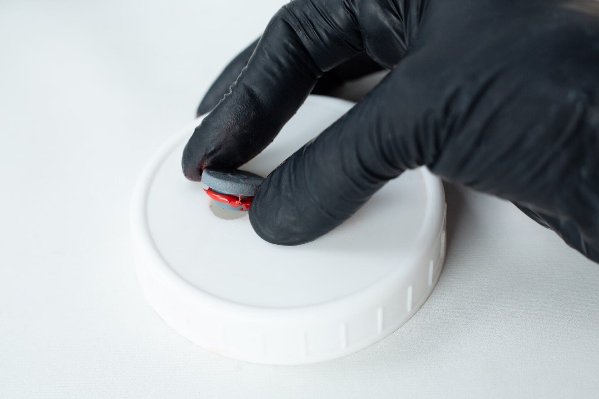 A gloved hand holds a small round object above a white plastic lid, ready to apply High Temperature RTV Silicone-Red on a light surface.
