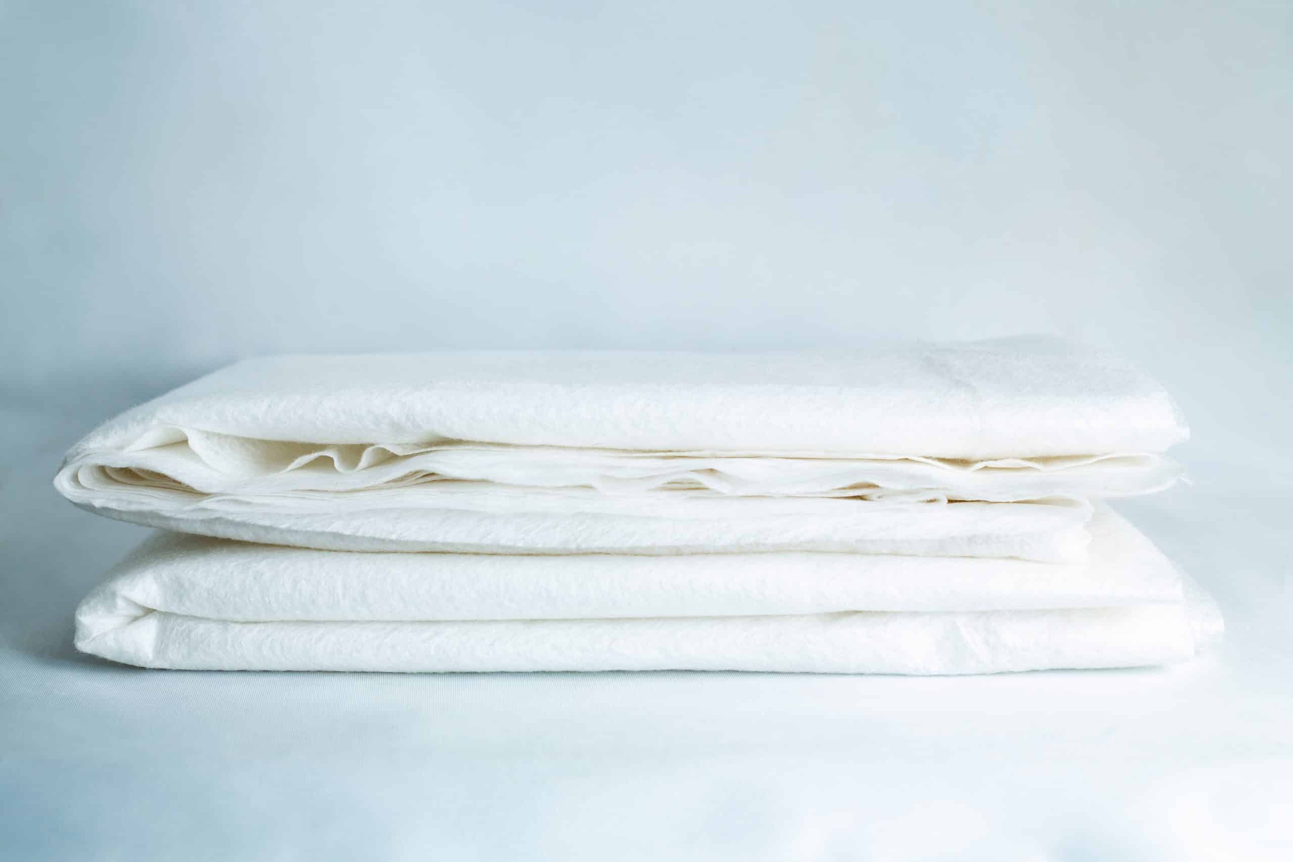 Two neatly folded white bed sheets rest on a light surface with a soft blue backdrop, making them an ideal match for the Light Log Blanket.
