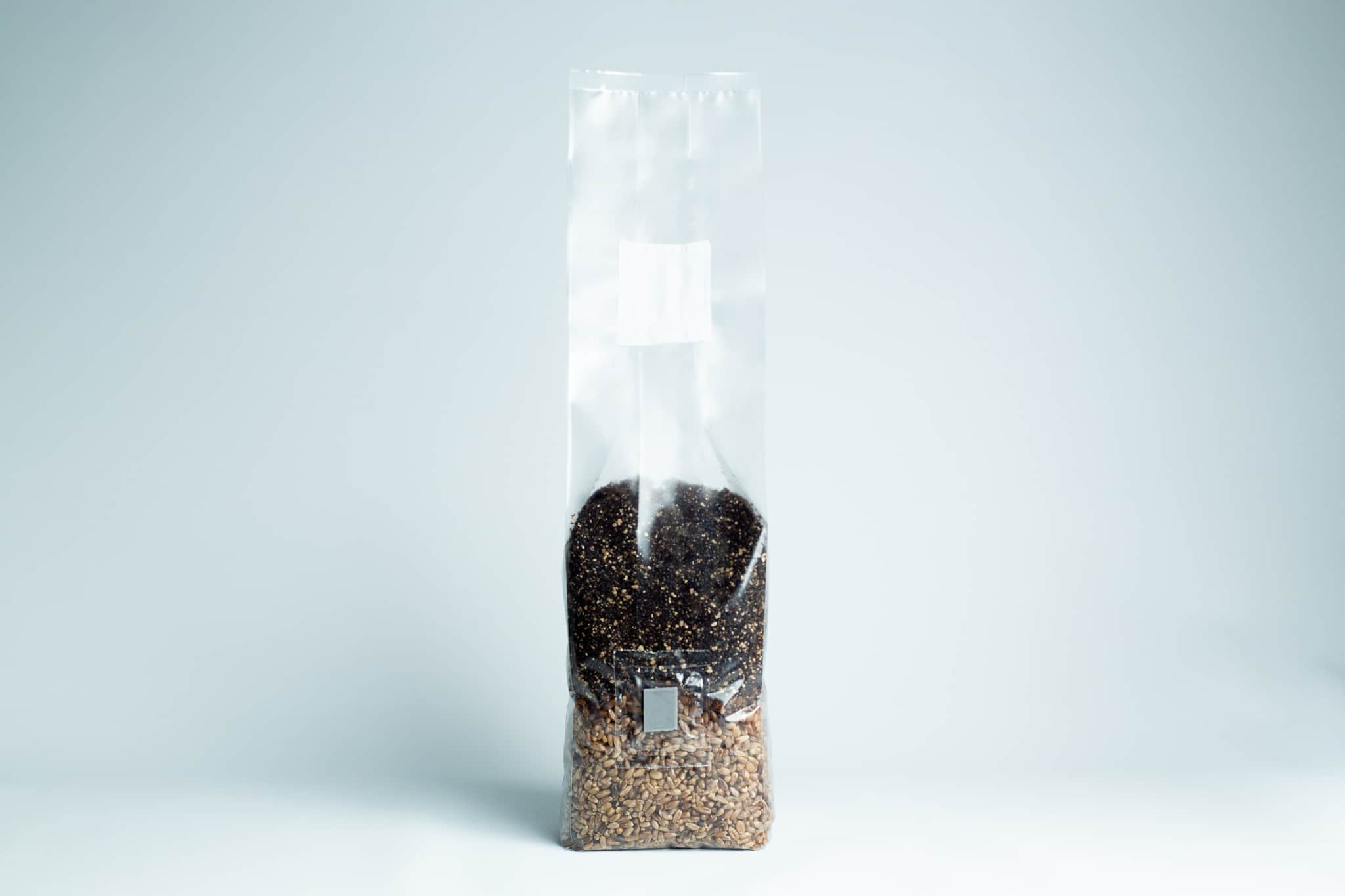 A clear plastic bag filled with brown and black seeds, standing upright on a light surface.