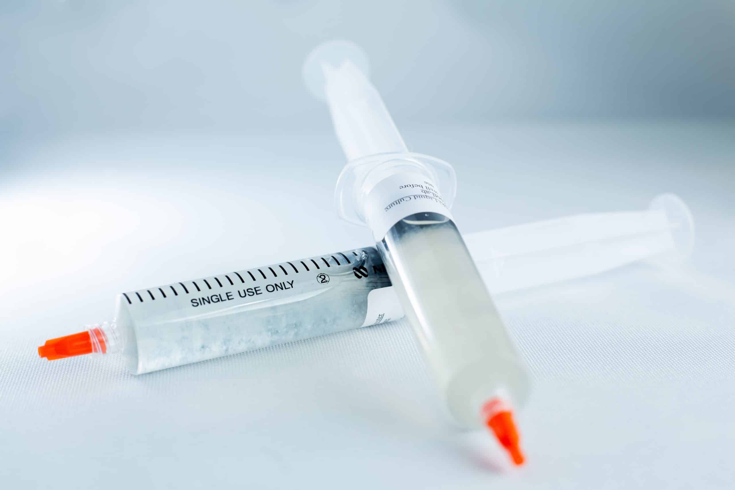 Two single-use syringes with orange caps containing Mushroom Liquid Culture | Wood Ear | Auricularia auricula-judae lie on a white surface, one filled with a clear liquid.