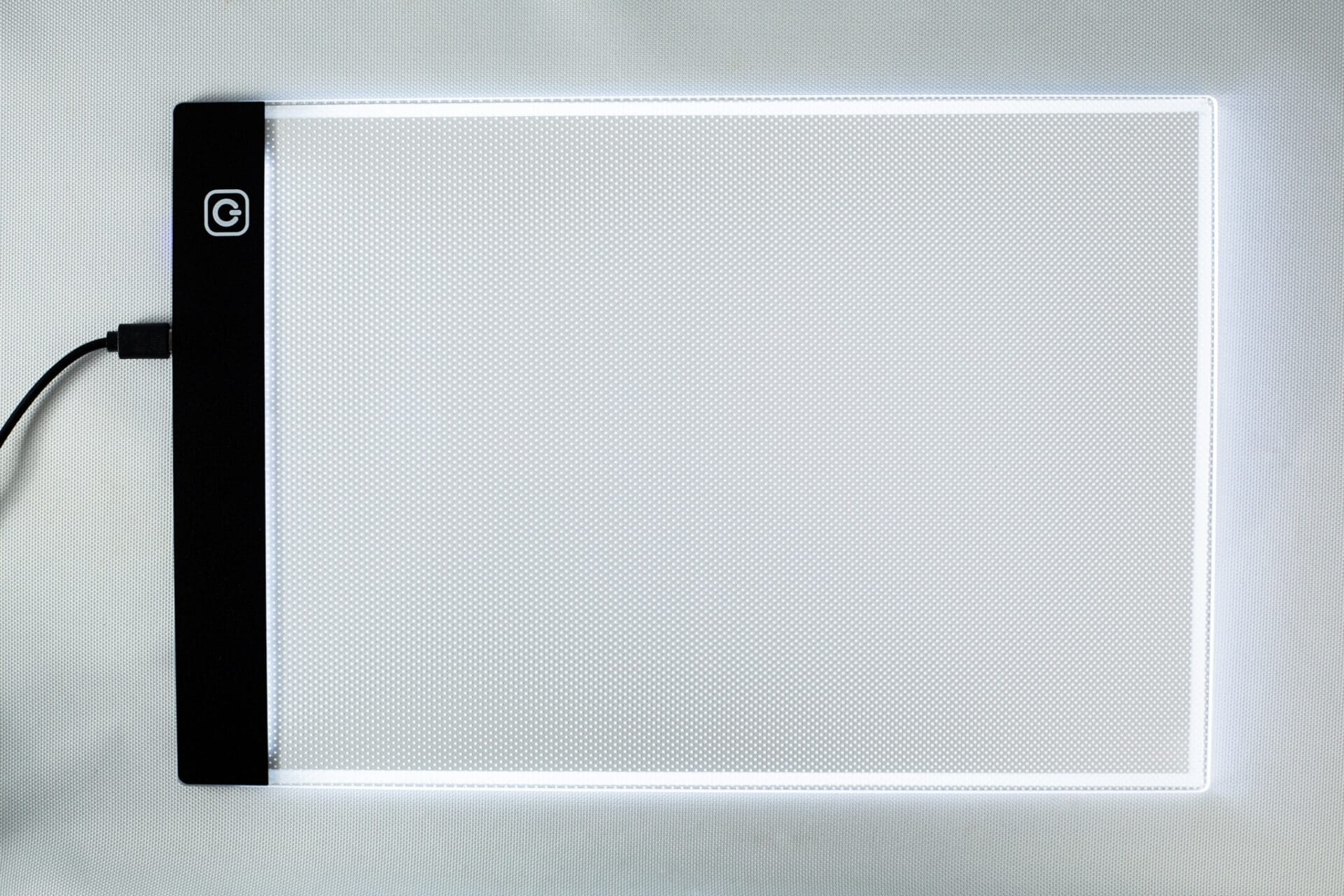 A rectangular LED light pad with a black edge and glowing white surface, plugged in on the left-hand side.