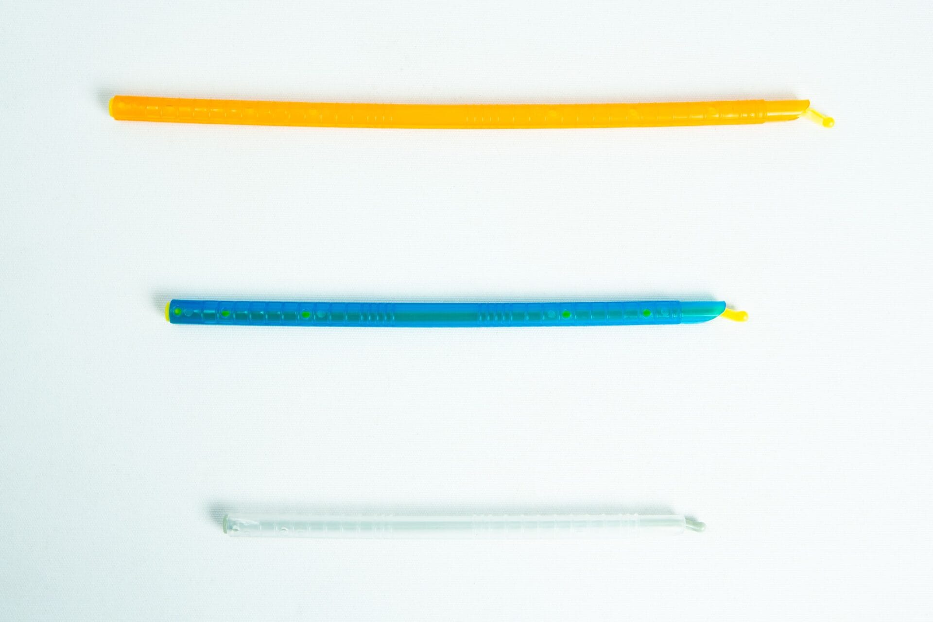 Three colourful twist ties, orange, blue, and clear, are laid out horizontally on a white background.