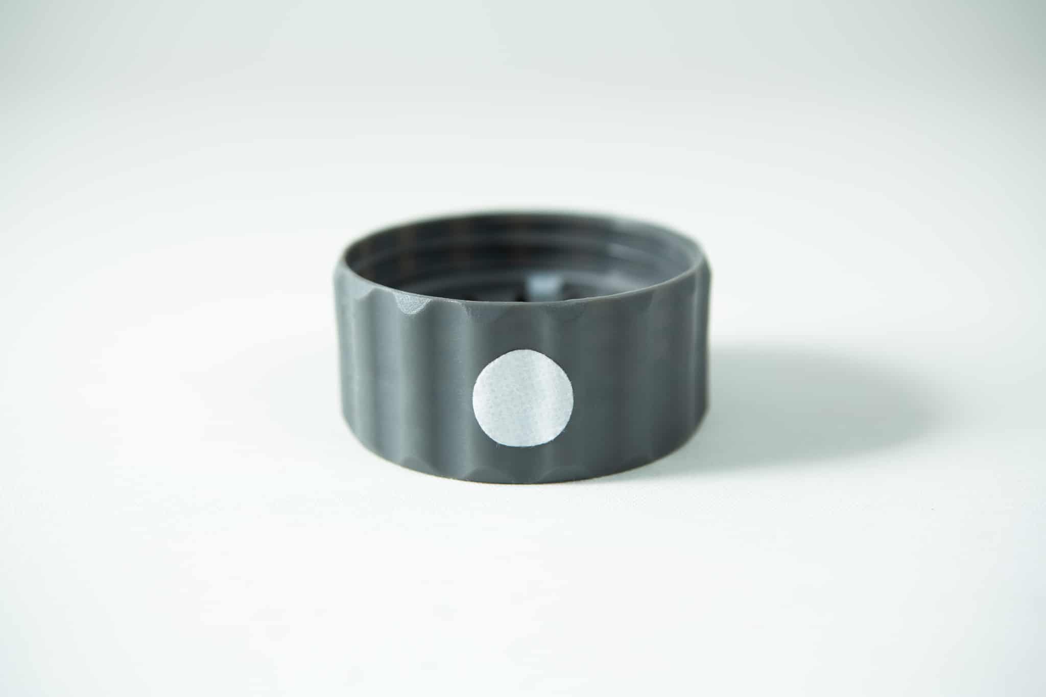 A Plant Cell Biocoupler™ Glass Set bottle cap—grey plastic with a white circular sticker on its side—rests on a white surface.